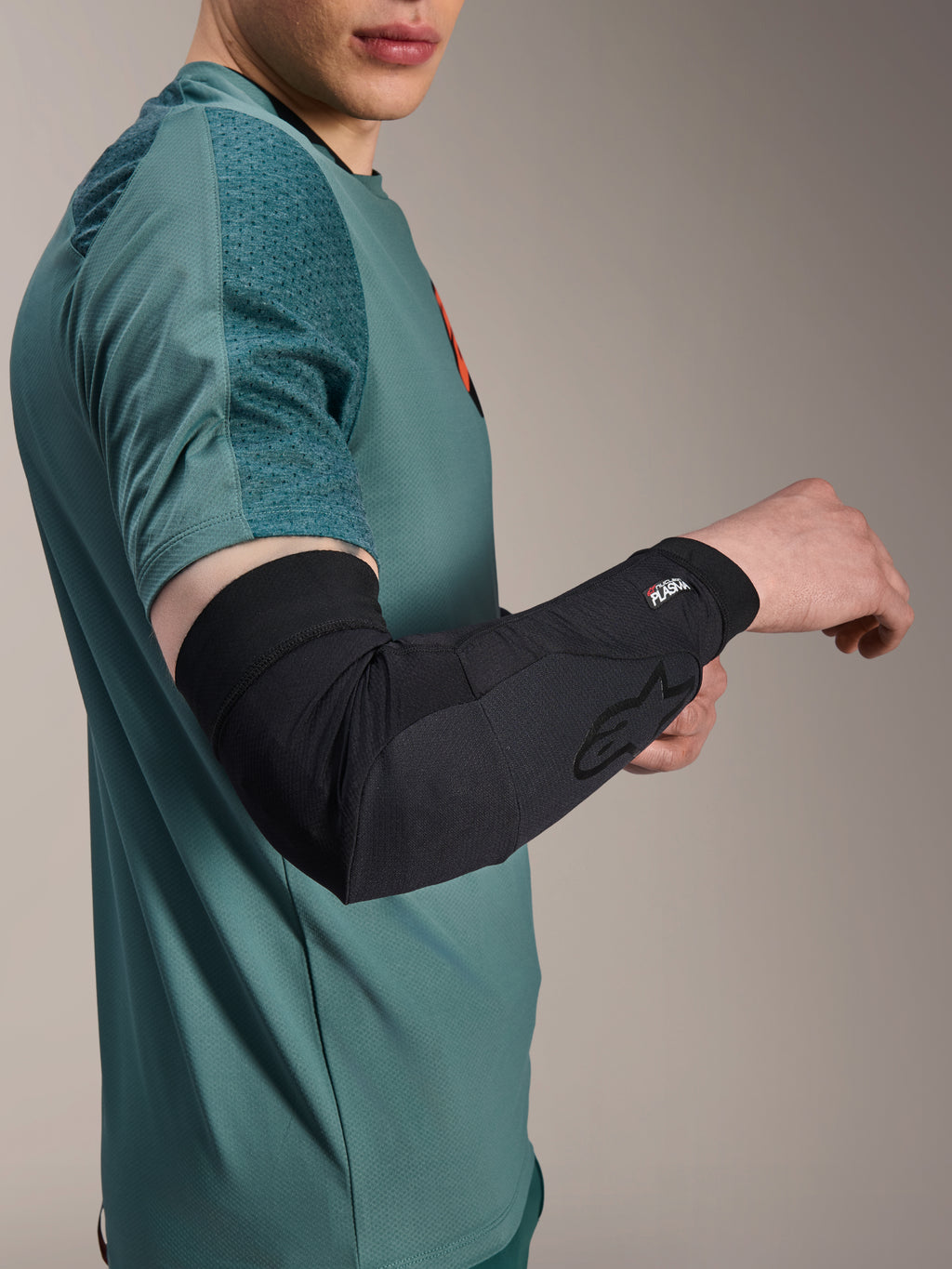 Alpinestars A-Motion Plasma elleboogmouw, fiets elleboogbeschermer, zwart, gedragen door een rijder met een groenblauw shirt, voorzien van Alpinestars-logo en Nucleon Plasma-branding, slank en ademend ontwerp voor MTB-bescherming