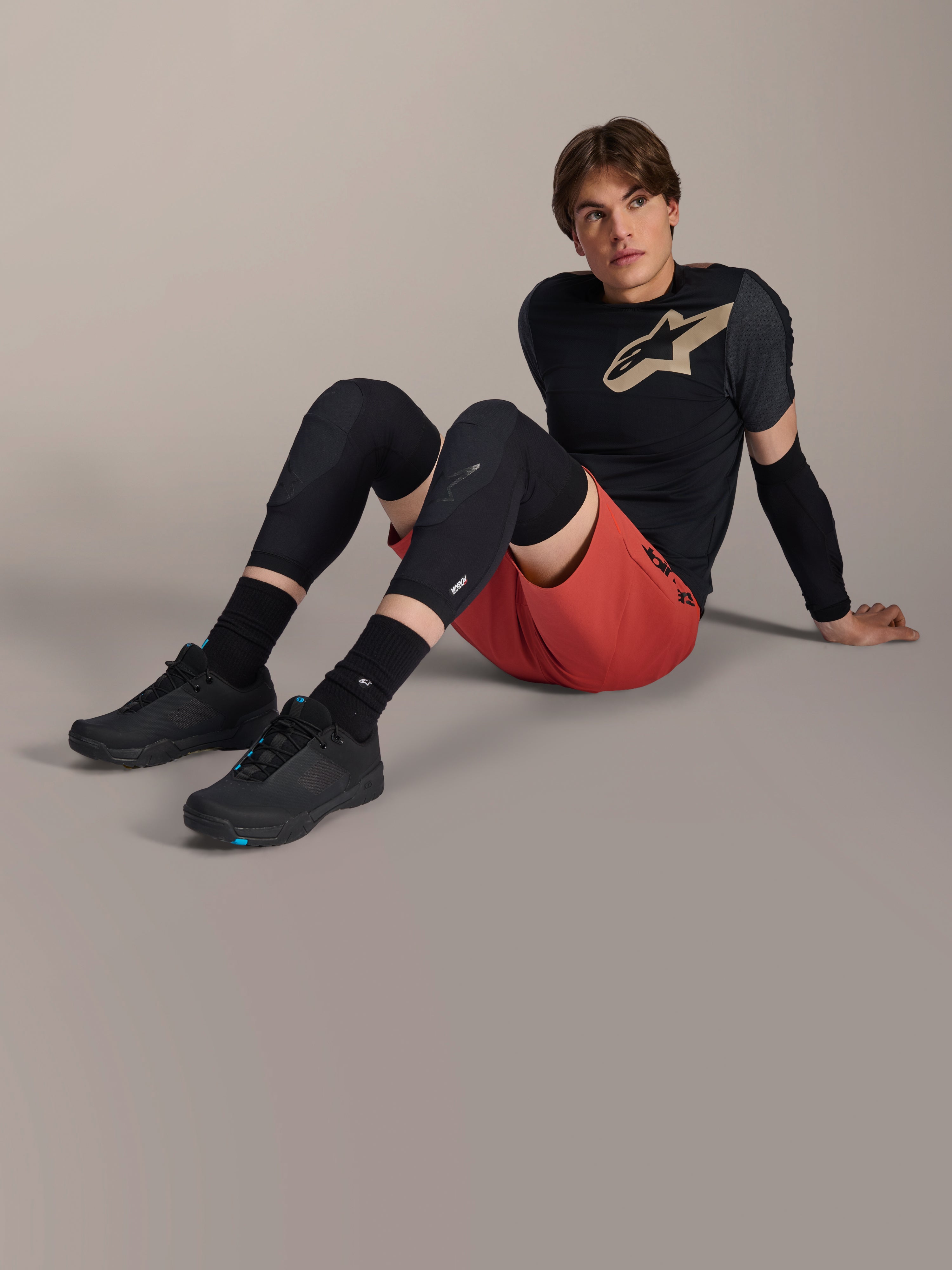 Alpinestars A-Motion Plasma elleboogbeschermer, gedragen door een mannelijke rijder zittend op een studiovloer, met zwarte mesh-constructie, gecombineerd met een rode short en een zwarte Alpinestars jersey met beige logo, hoogwaardige mountainbikebescherming voor trailrijden.