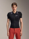 Male model wearing Alpinestars A-Motion Plasma Protection Jacket - Short Sleeve, Black with gray camo shoulder padding, white Alpinestars logo on the chest, paired with red pants, slim-fit MTB body armor with a front zipper