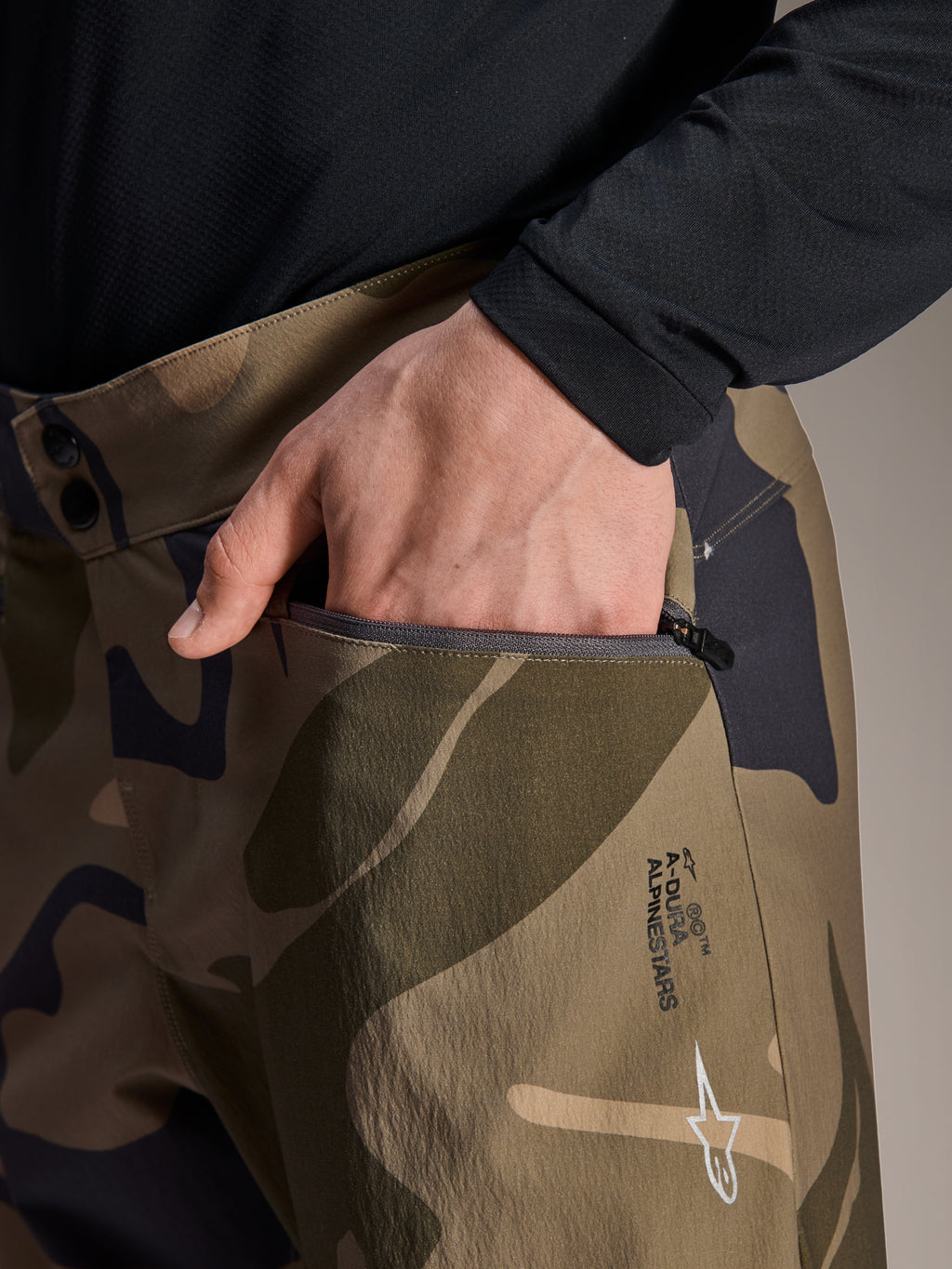 Close-up detail van Alpinestars A-Dura Camo Pant, bruine camouflage mountainbikebroek, met een hand in een zak met ritssluiting, dubbele drukknoopsluiting in de taille en Alpinestars-branding op het dijbeen.