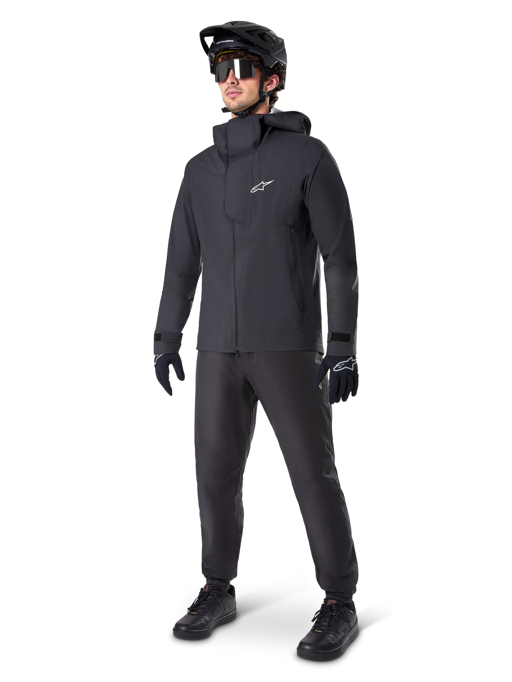 Mannelijke mountainbiker in zwarte Alpinestars A-Dura Waterdichte Broek, bijpassend zwart waterdicht jack met capuchon, zwarte MTB-helm met vizier, donkere fietsbril en zwarte handschoenen, complete professionele uitrusting voor trailrijden in alle weersomstandigheden