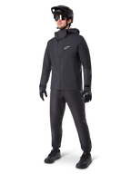 Mannelijke mountainbiker in zwarte Alpinestars A-Dura Waterdichte Broek, bijpassend zwart waterdicht jack met capuchon, zwarte MTB-helm met vizier, donkere fietsbril en zwarte handschoenen, complete professionele uitrusting voor trailrijden in alle weersomstandigheden