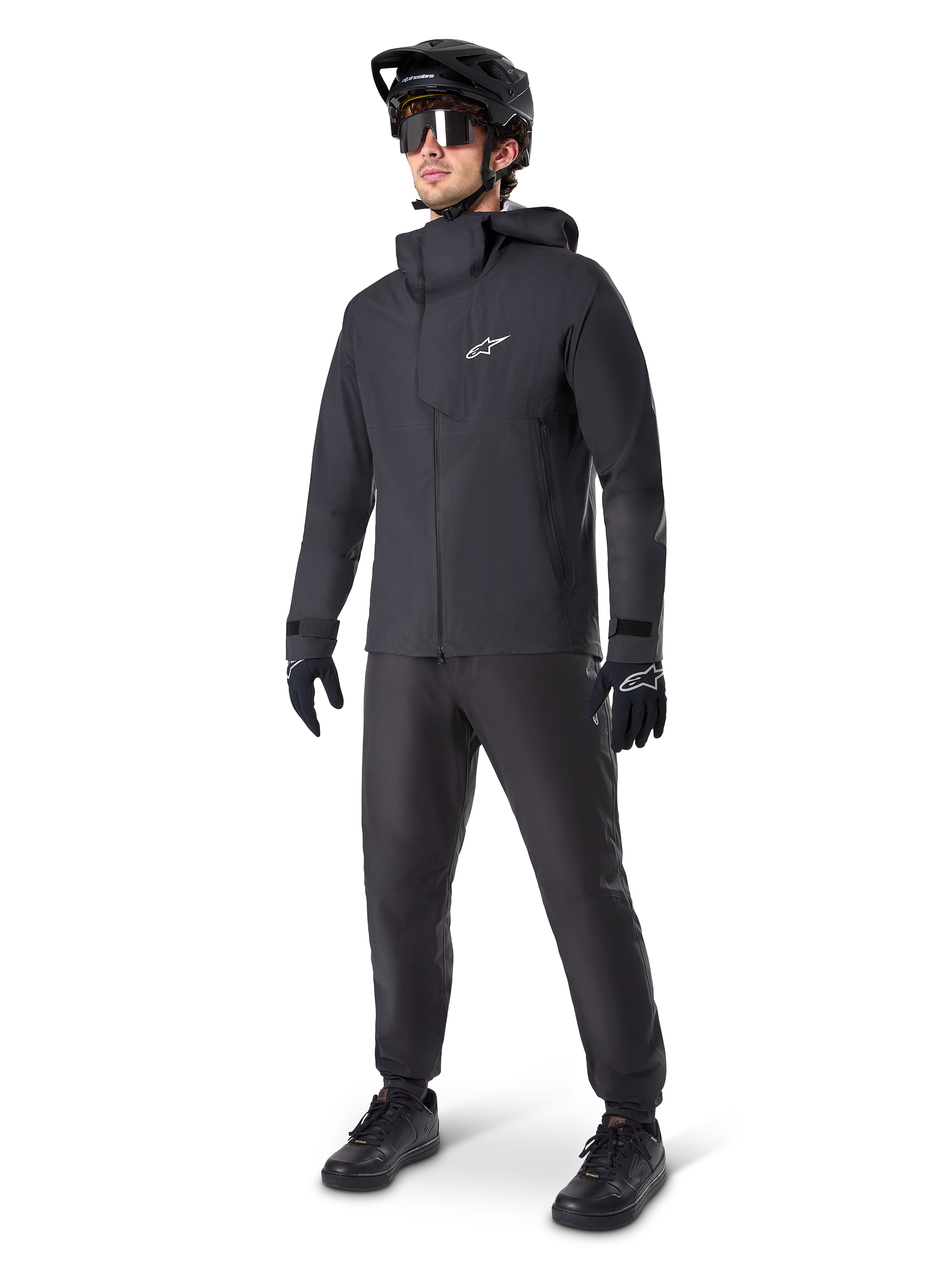 Mannelijke mountainbiker in zwarte Alpinestars A-Dura Waterdichte Broek, bijpassend zwart waterdicht jack met capuchon, zwarte MTB-helm met vizier, donkere fietsbril en zwarte handschoenen, complete professionele uitrusting voor trailrijden in alle weersomstandigheden