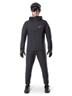 Alpinestars A-Dura Waterproof Pants, zwarte MTB-broek, gedragen als onderdeel van een complete volledig zwarte mountainbike-uitrusting inclusief een technisch waterdicht jack, zwarte helm, zonnebril en handschoenen, ontworpen voor off-road ritten in natte weersomstandigheden.