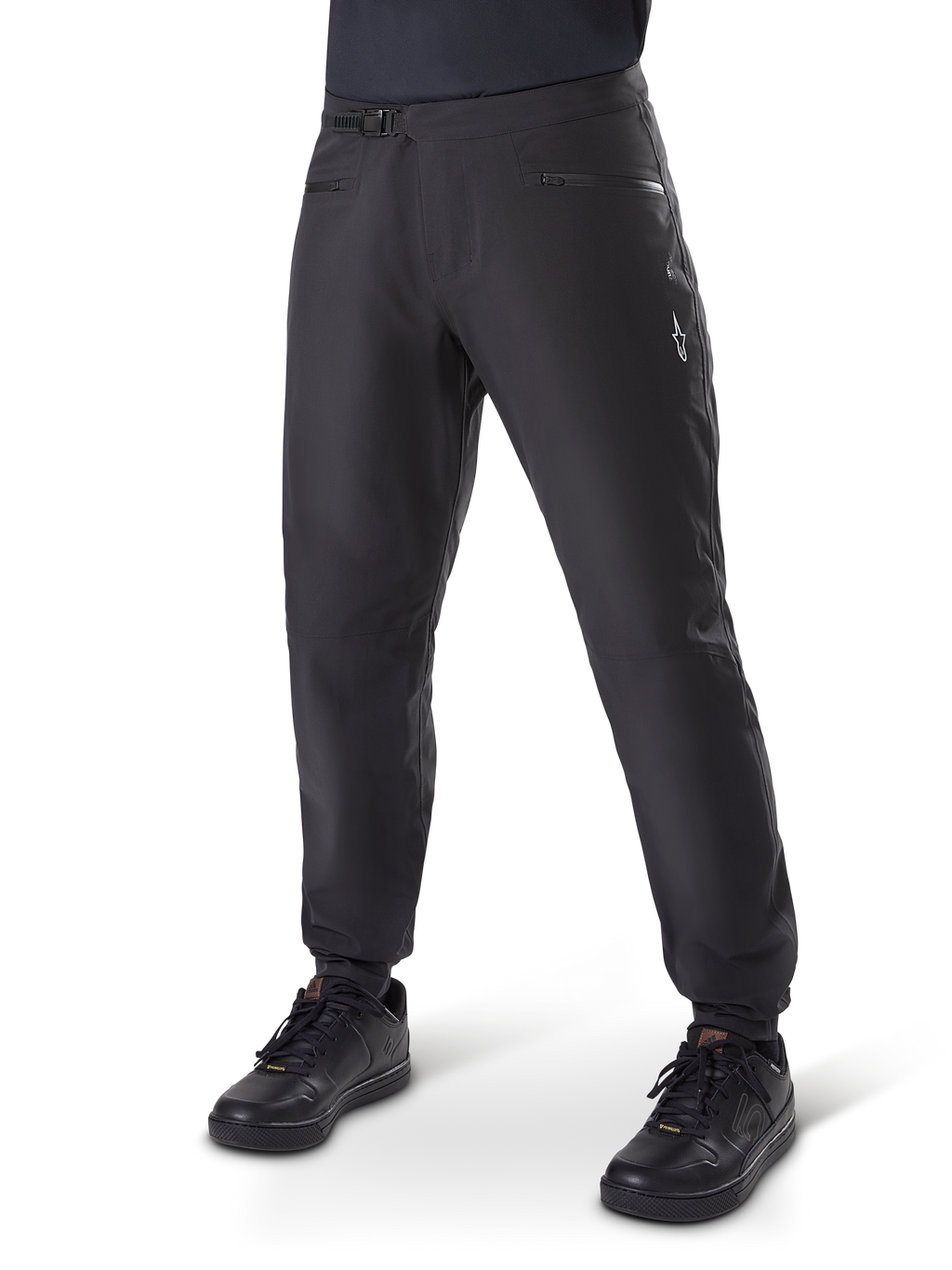Alpinestars A-Dura Waterproof Pants, MTB-broek, zwart, voorzien van een ratelsluiting bij de taille, zakken met ritssluiting en een wit Astars-logo op de dij, gedragen met zwarte fietsschoenen voor trailrijden in alle weersomstandigheden.