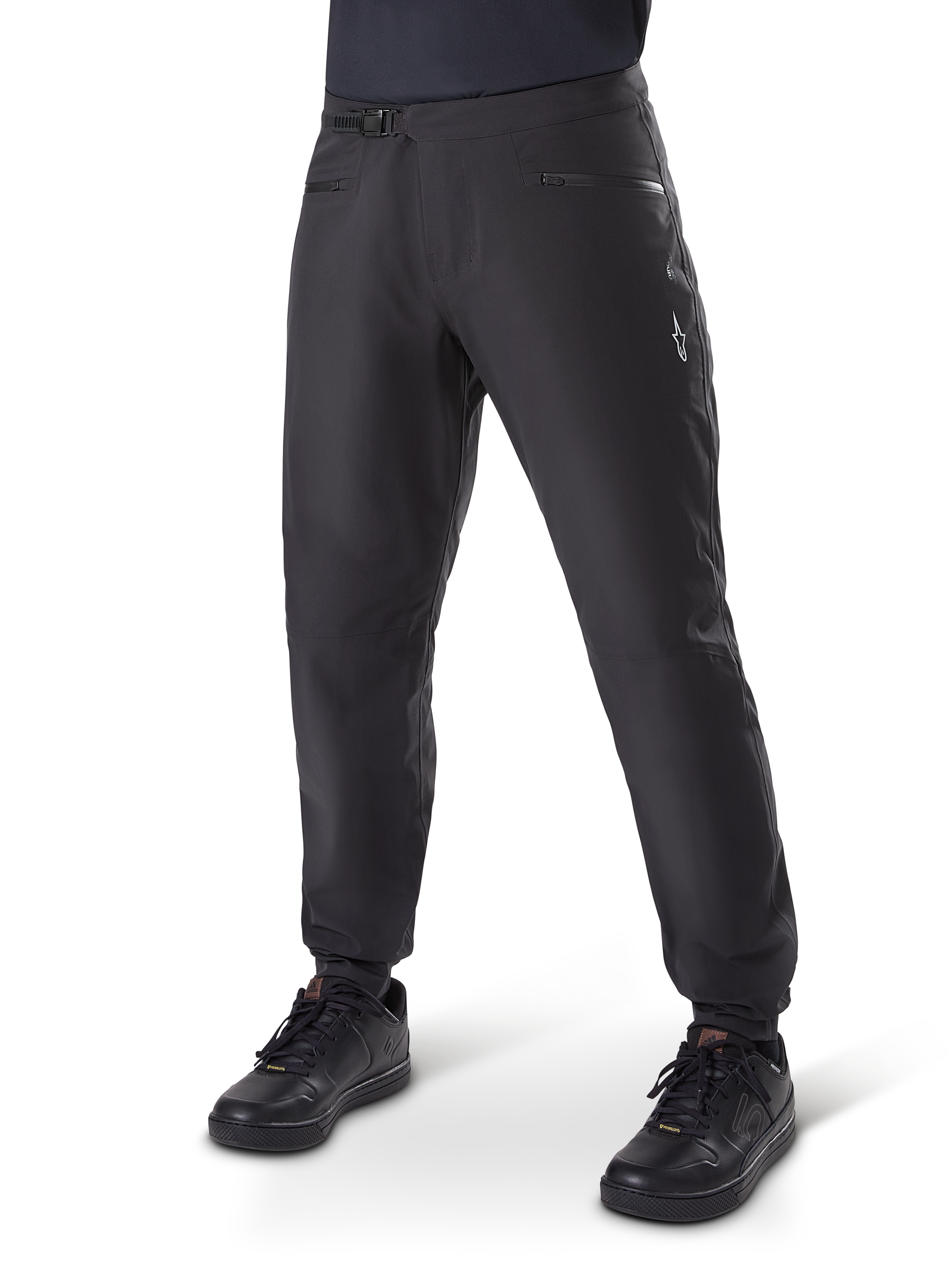 Alpinestars A-Dura Waterproof Pants, MTB-broek, zwart, voorzien van een ratelsluiting bij de taille, zakken met ritssluiting en een wit Astars-logo op de dij, gedragen met zwarte fietsschoenen voor trailrijden in alle weersomstandigheden.