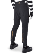 Alpinestars A-Aria Elite broek, zwarte MTB-broek, achteraanzicht, met verticale gouden Alpinestars-branding op de achterkant van de broekspijpen, gedragen met een zwart-wit gestreept shirt en zwarte schoenen, technische fietskleding voor mountainbiken