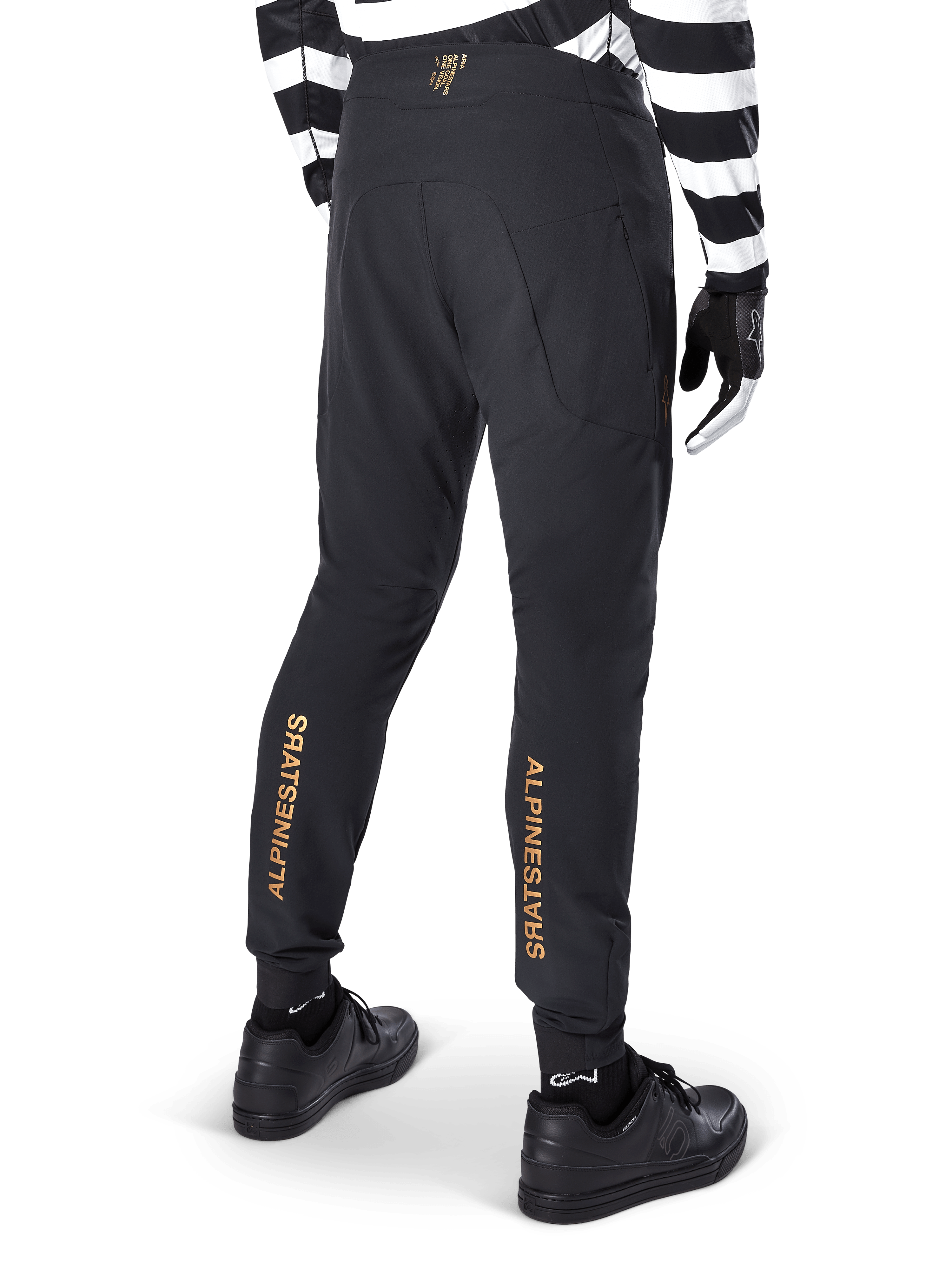 Alpinestars A-Aria Elite broek, zwarte MTB-broek, achteraanzicht, met verticale gouden Alpinestars-branding op de achterkant van de broekspijpen, gedragen met een zwart-wit gestreept shirt en zwarte schoenen, technische fietskleding voor mountainbiken