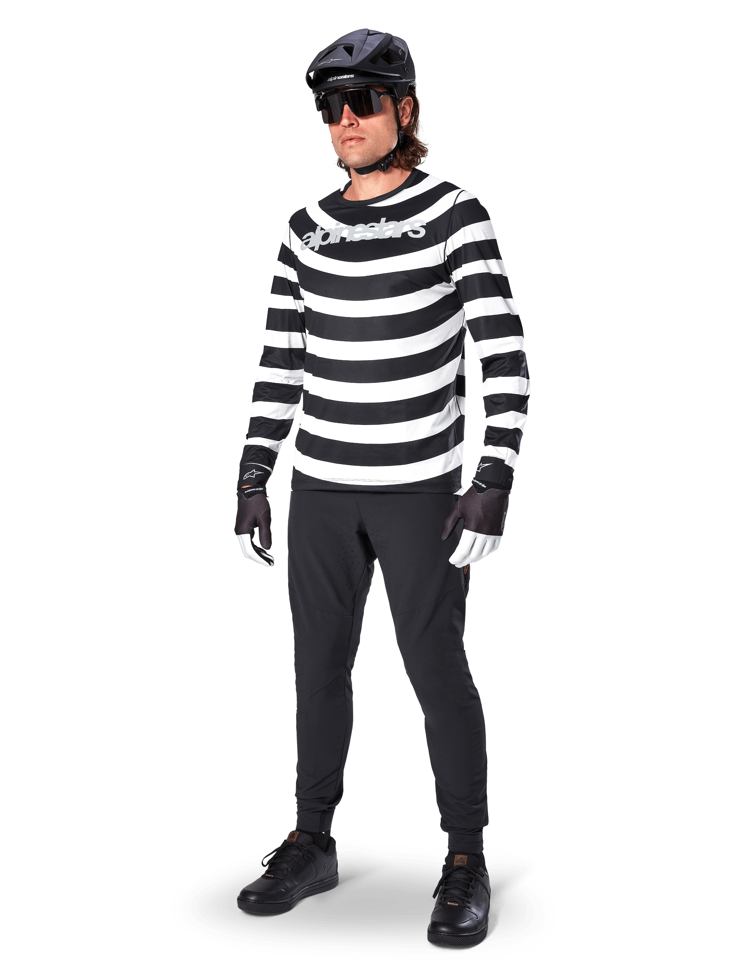 Alpinestars A-Aria Elite broek, zwarte MTB-broek, gedragen door een rijder met een zwart-wit gestreept shirt met lange mouwen, zwarte helm, zonnebril en zwart-witte handschoenen, performancekleding voor off-road fietsen