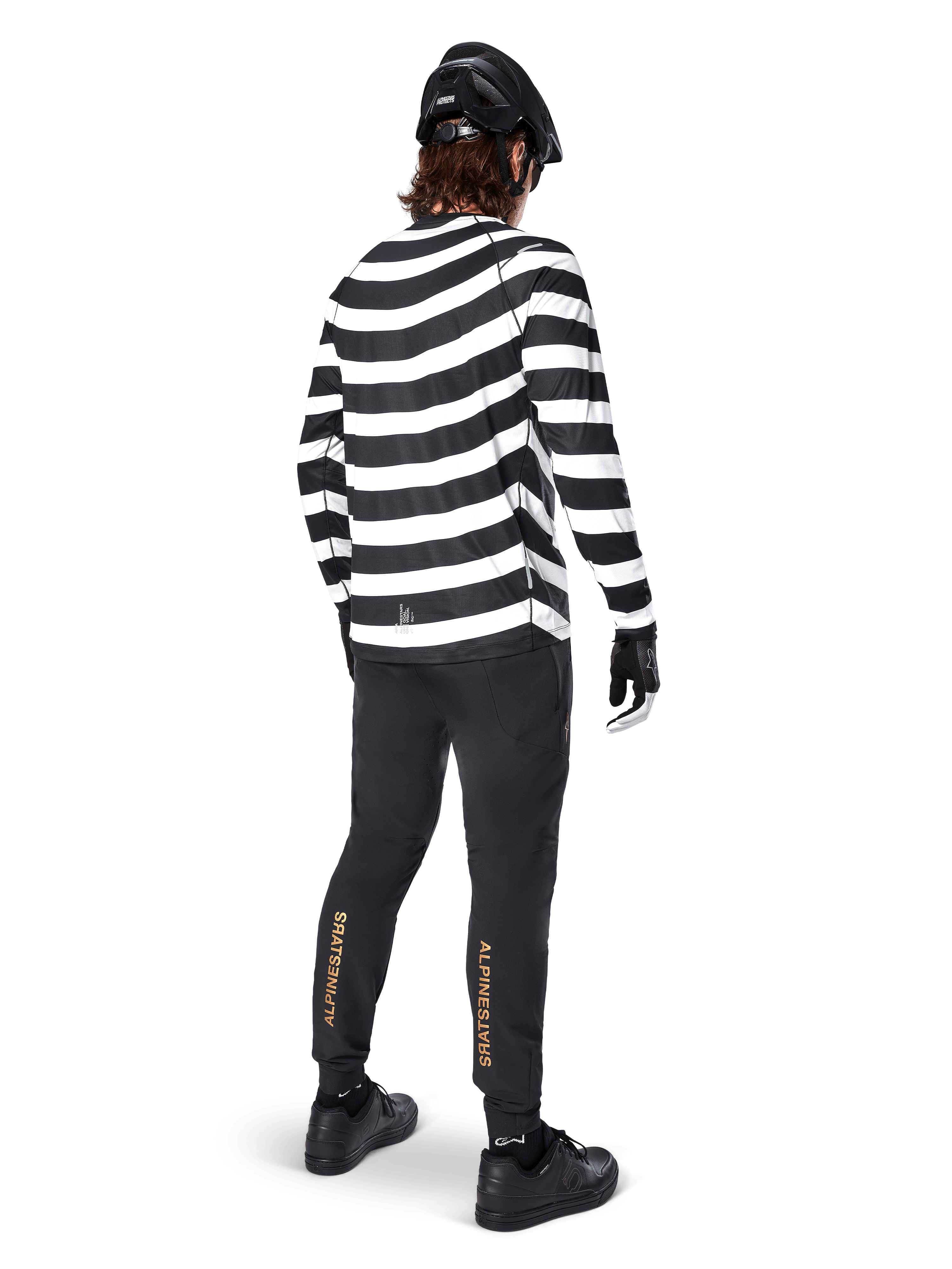 Alpinestars A-Aria Elite Pants, zwarte MTB-broek, met verticaal goudkleurig logo op de kuiten, gedragen door een rijder met een zwart-wit gestreepte jersey met lange mouwen, zwarte helm en zwarte schoenen, achteraanzicht toont de prestatiegerichte pasvorm en het trail-ready ontwerp