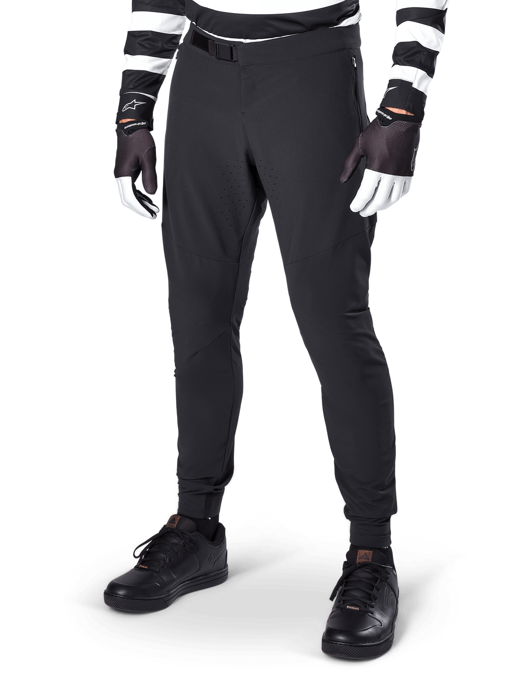 Alpinestars A-Aria Elite broek, zwarte mountainbikebroek, met lasergesneden ventilatie op de dijen en magnetische gespsluiting, gedragen met zwart-wit gestreepte jersey en handschoenen, performance MTB-kleding voor off-road fietsen