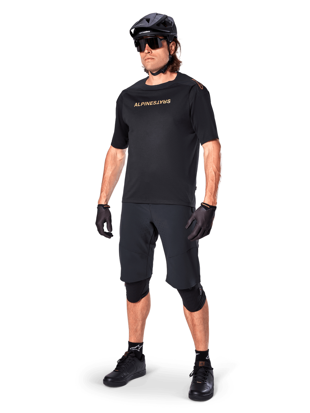Fietser die Alpinestars A-Aria Elite shorts draagt, zwarte mountainbikebroek, gecombineerd met een bijpassende zwarte jersey met gouden branding, zwarte helm, zonnebril en kniebeschermers, complete performance-outfit voor off-road fietsen