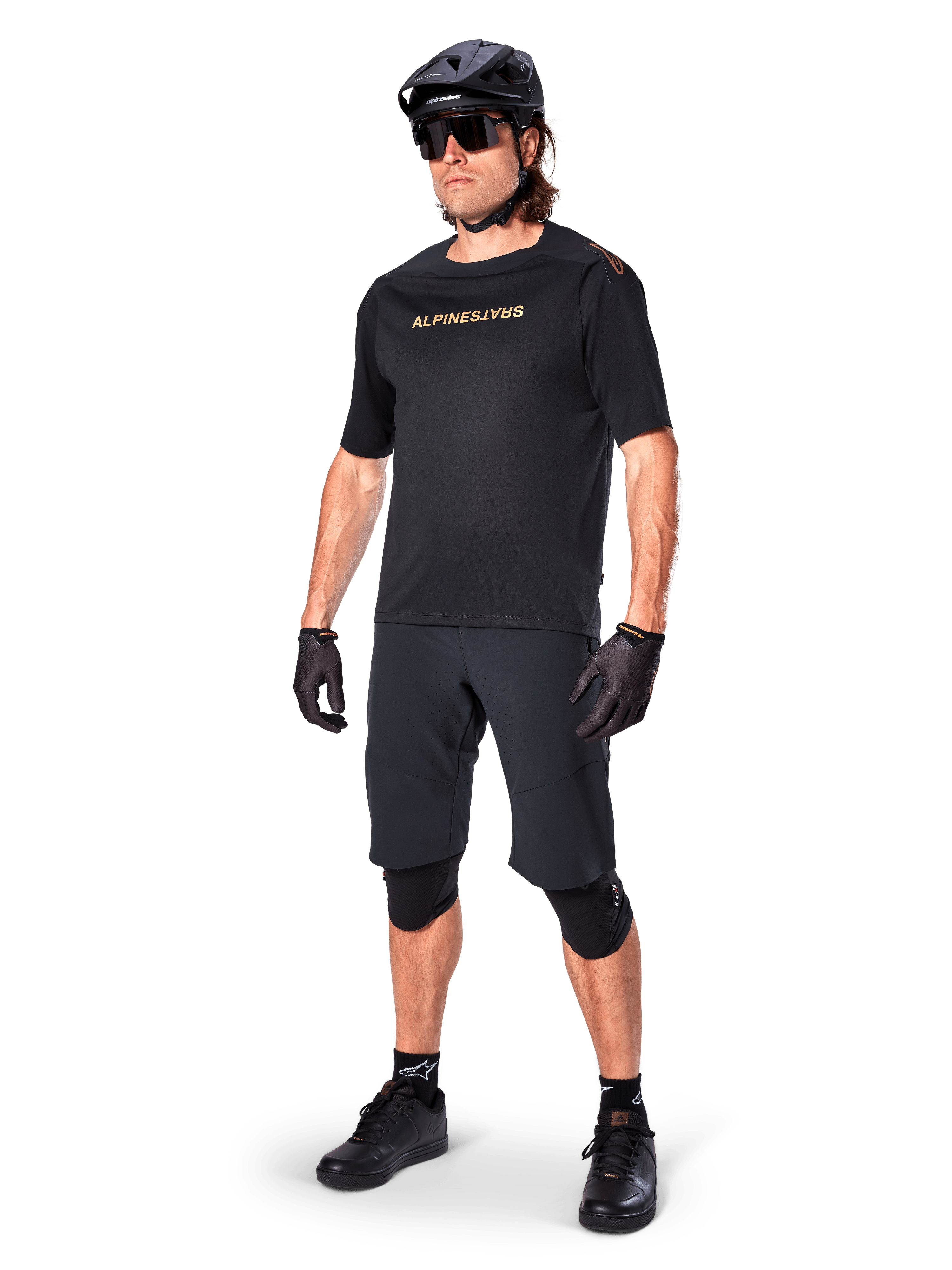 Fietser die Alpinestars A-Aria Elite shorts draagt, zwarte mountainbikebroek, gecombineerd met een bijpassende zwarte jersey met gouden branding, zwarte helm, zonnebril en kniebeschermers, complete performance-outfit voor off-road fietsen