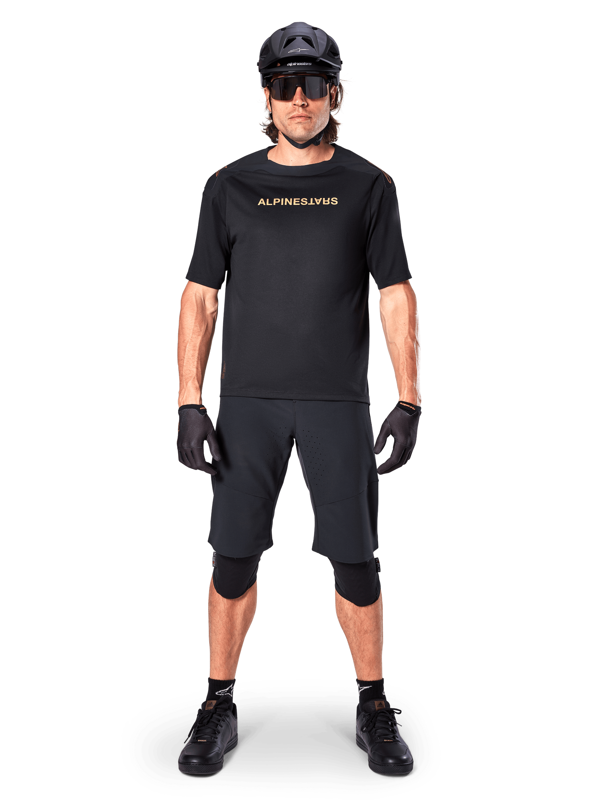 Mannelijke mountainbiker die zwarte Alpinestars A-Aria Elite Shorts draagt, gecombineerd met een bijpassend zwart Alpinestars fietsshirt met korte mouwen, zwarte helm, handschoenen en kniebeschermers, complete off-road wieleroutfit