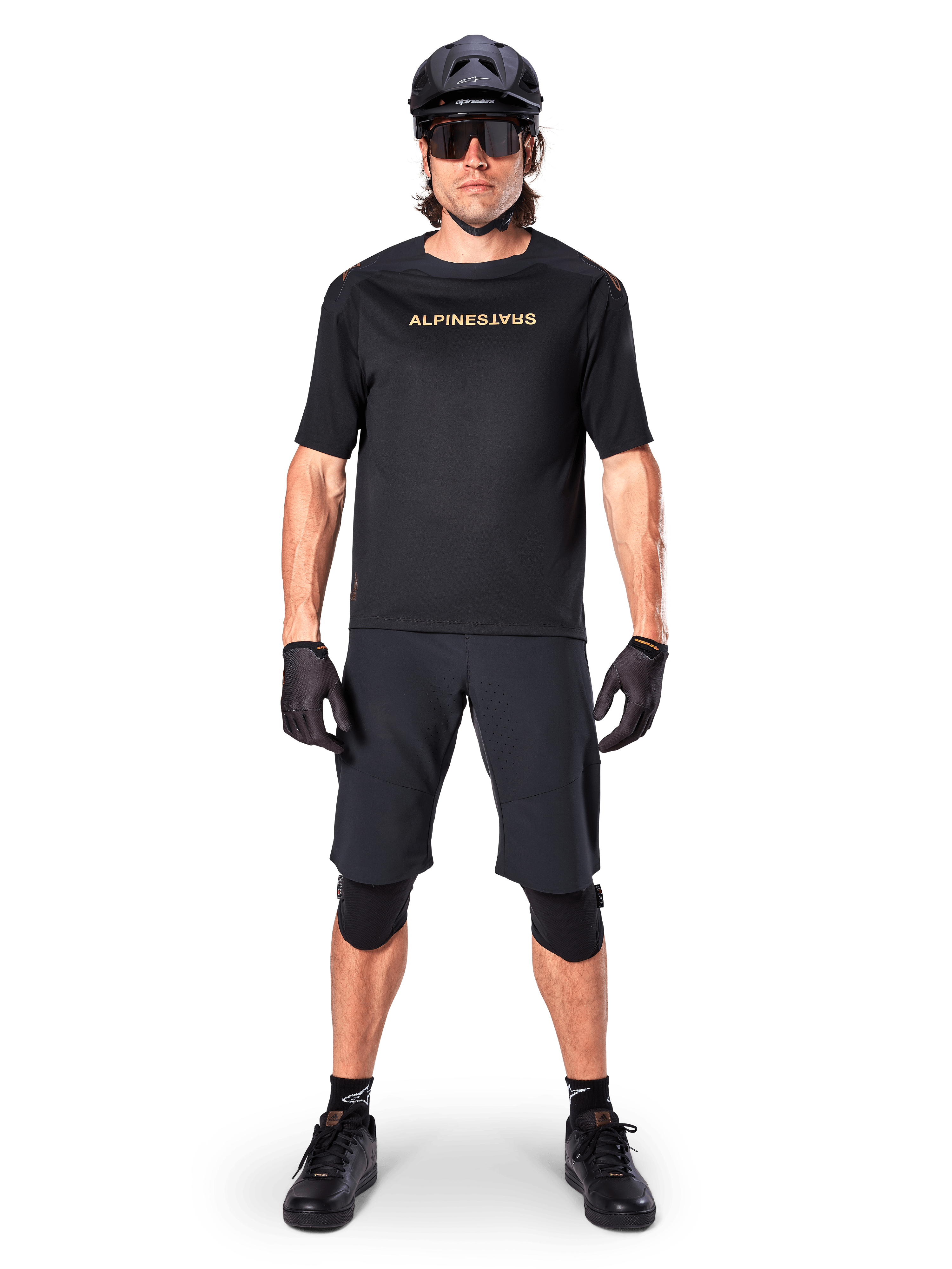 Mannelijke mountainbiker die zwarte Alpinestars A-Aria Elite Shorts draagt, gecombineerd met een bijpassend zwart Alpinestars fietsshirt met korte mouwen, zwarte helm, handschoenen en kniebeschermers, complete off-road wieleroutfit