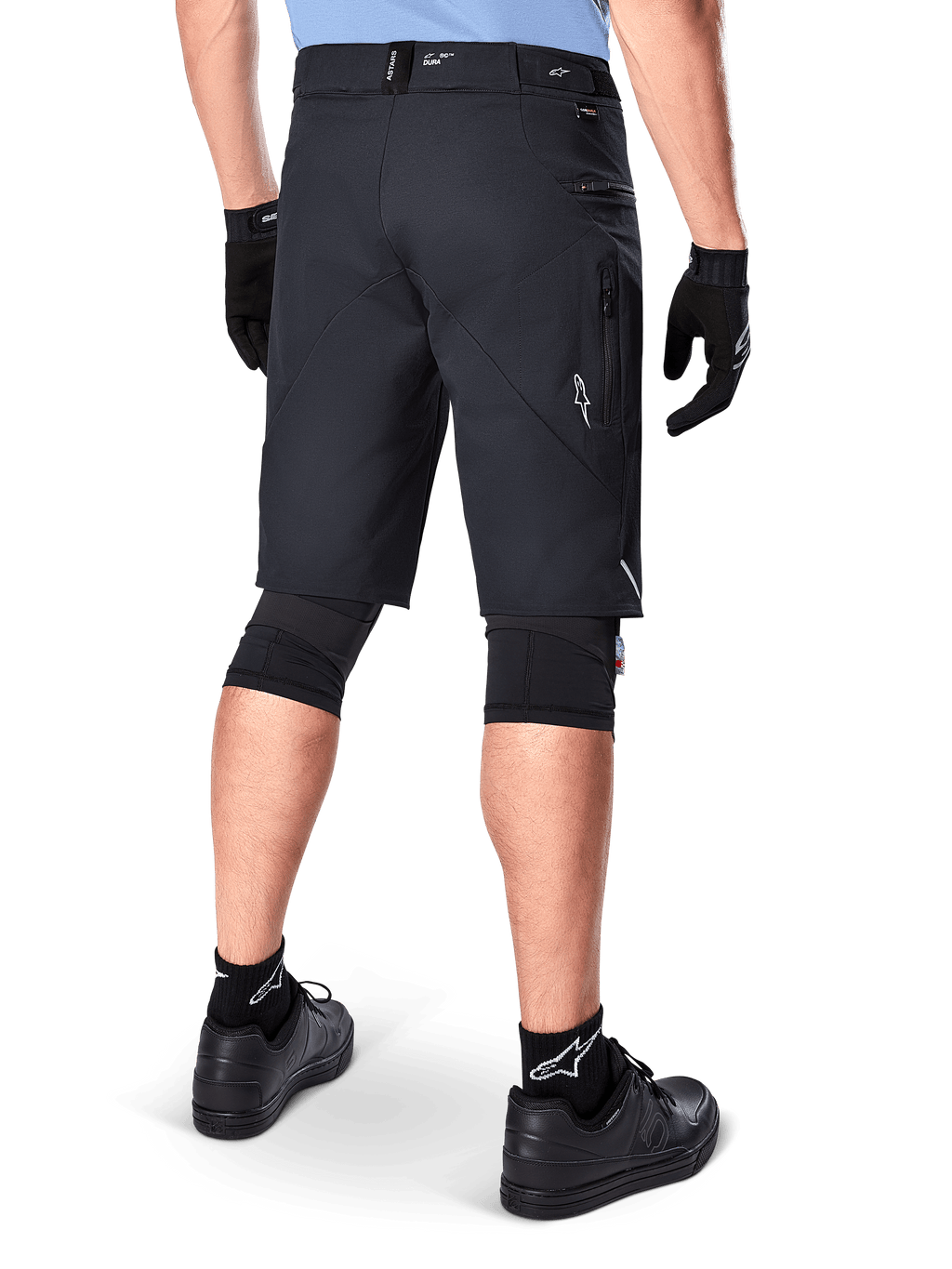 Alpinestars A-Dura Elite Shorts, zwart, achteraanzicht van een rijder in mountainbike-uitrusting inclusief zwarte kniebeschermers, handschoenen en sokken met witte logo's, voorzien van zijzakken met ritssluiting en reflecterende accenten op duurzame Cordura-stof.