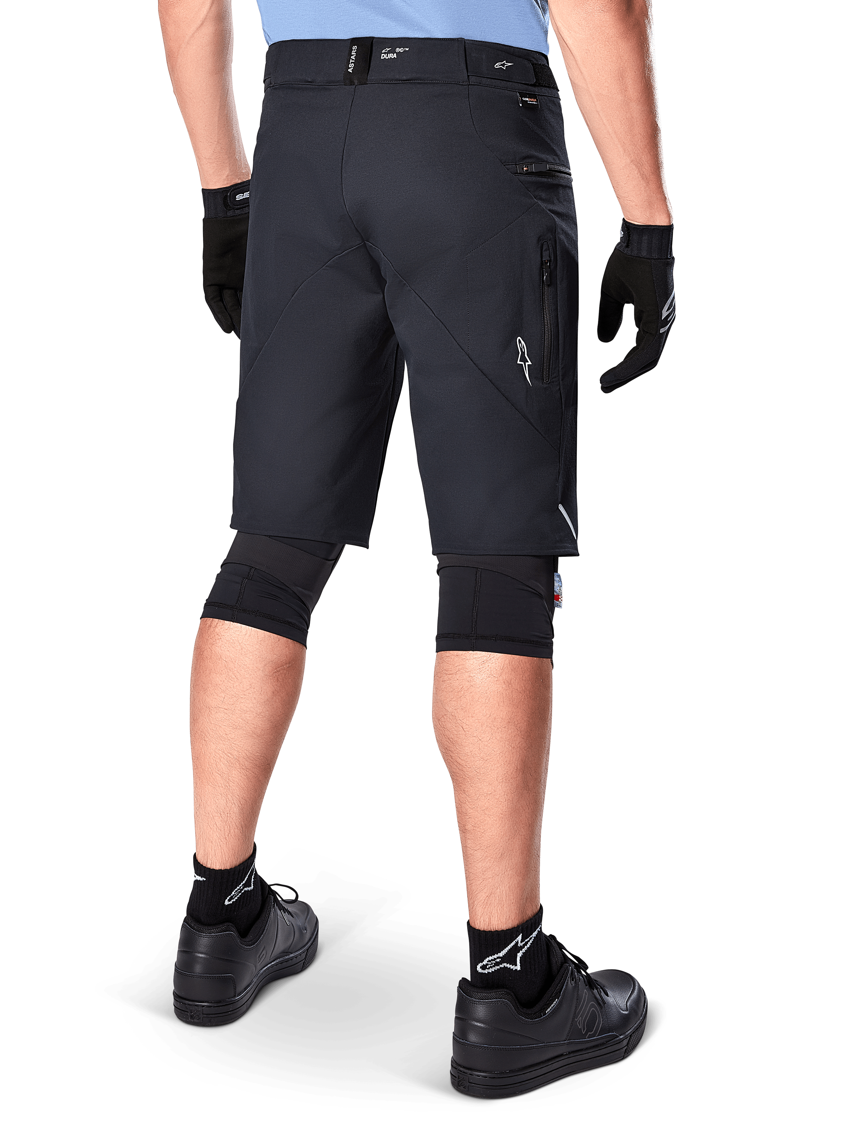 Alpinestars A-Dura Elite Shorts, zwart, achteraanzicht van een rijder in mountainbike-uitrusting inclusief zwarte kniebeschermers, handschoenen en sokken met witte logo's, voorzien van zijzakken met ritssluiting en reflecterende accenten op duurzame Cordura-stof.