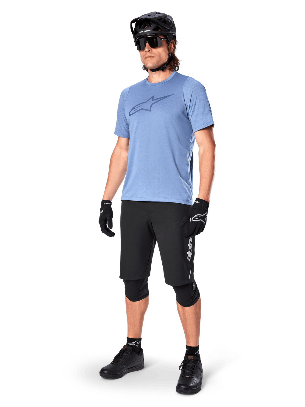 Mannelijke rijder in zwarte Alpinestars A-Dura Elite Shorts, lichtblauwe jersey met korte mouwen en logo op de borst, zwarte helm, zonnebril en beschermende kniebeschermers, volledige mountainbike-uitrusting voor off-road trails