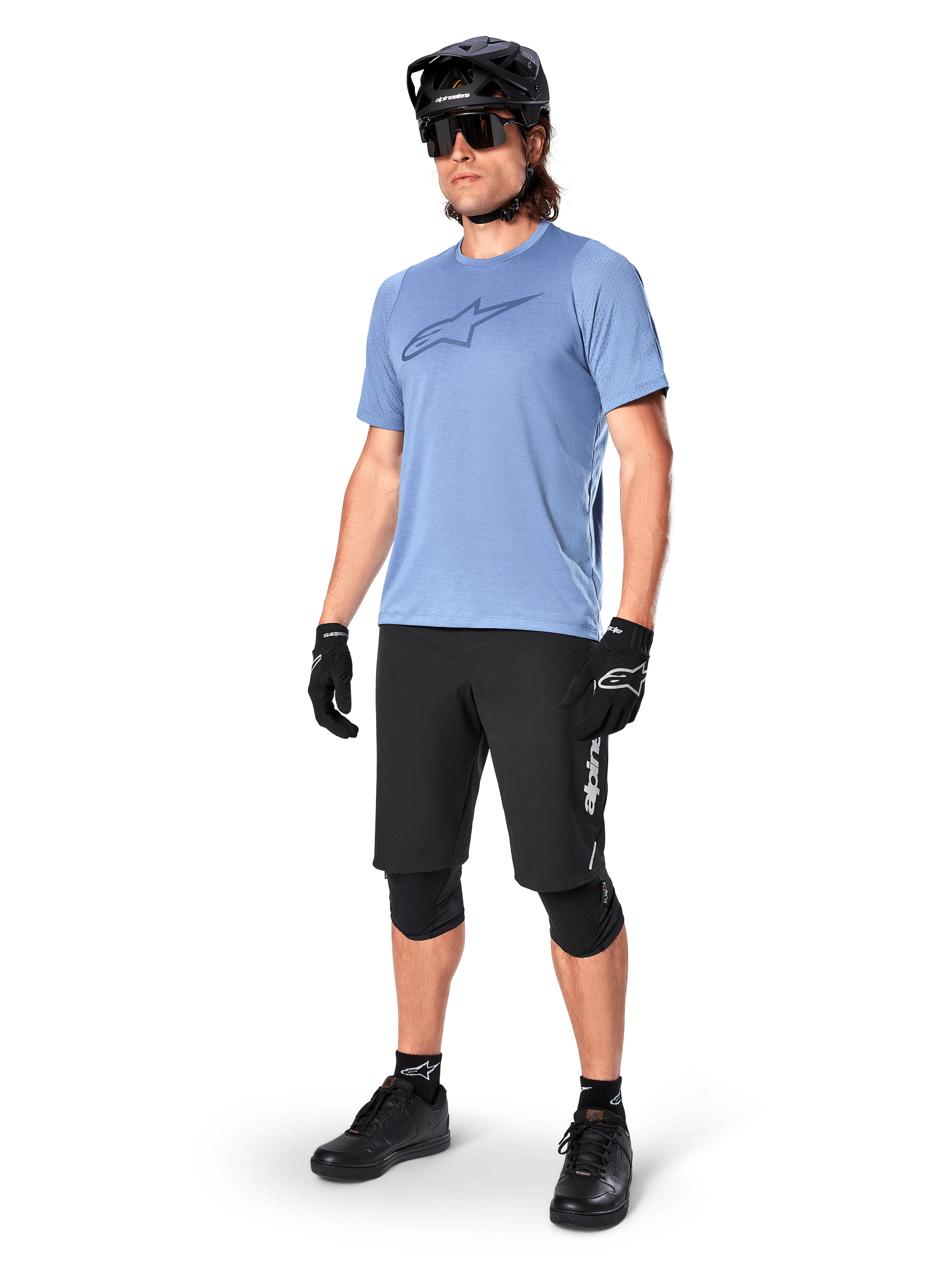 Mannelijke rijder in zwarte Alpinestars A-Dura Elite Shorts, lichtblauwe jersey met korte mouwen en logo op de borst, zwarte helm, zonnebril en beschermende kniebeschermers, volledige mountainbike-uitrusting voor off-road trails