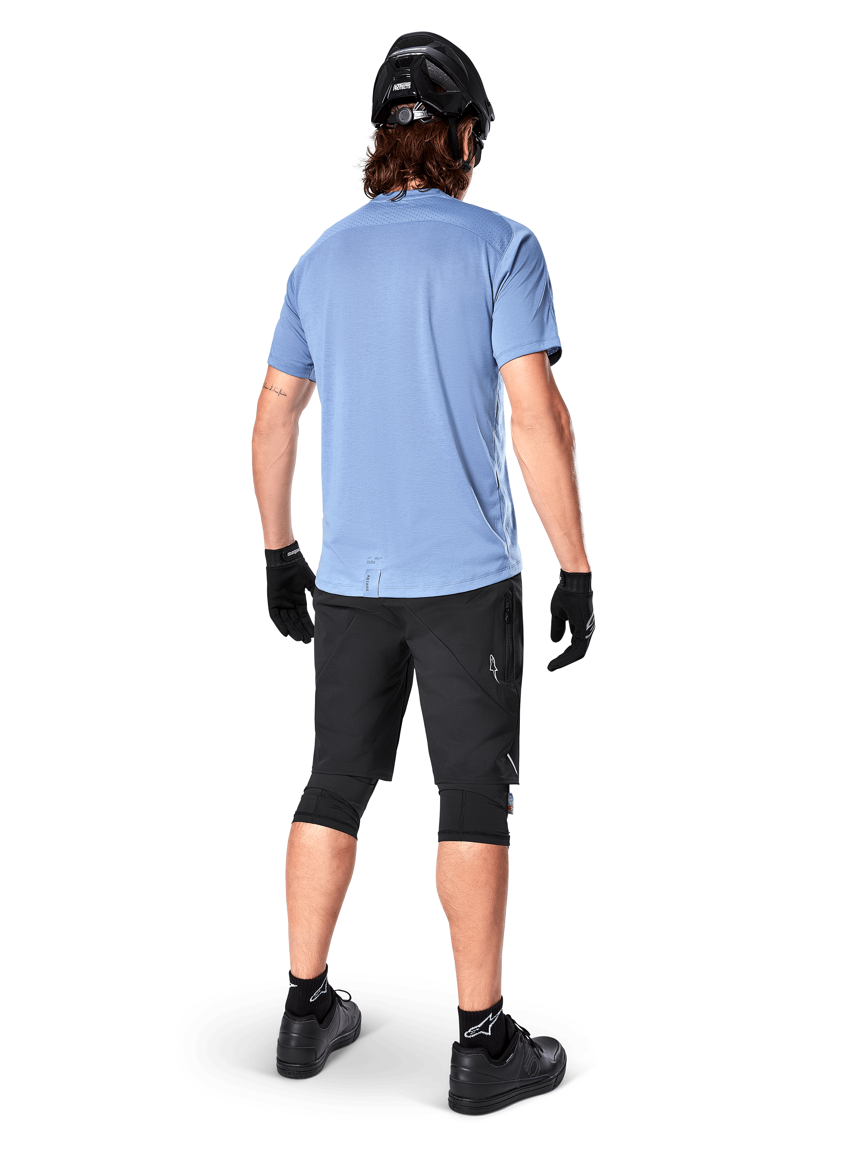 Alpinestars A-Dura Elite Shorts, mountainbikebroek, zwart, achteraanzicht op model, gedragen met lichtblauw shirt met korte mouwen en zwarte beschermende uitrusting, technisch ontwerp met ritszakken en reflecterende details voor off-road fietsen