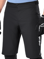 Alpinestars A-Dura Elite Shorts, zwarte MTB-shorts, close-up met drukknoopsluiting aan de voorzijde en zijzak met rits, gedragen door een rijder met zwarte fietshandschoenen en kniebeschermers, performance gear voor mountainbiken