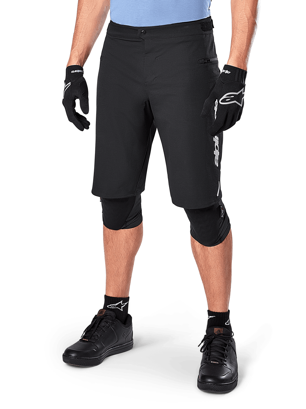 Alpinestars A-Dura Elite Shorts, zwarte MTB-mountainbikeshorts, gedragen met een lichtblauw shirt en zwarte kniebescherming, voorzien van een stevige drukknoopsluiting en een zak met rits, gecombineerd met zwarte fietsschoenen en handschoenen voor een complete off-road outfit.