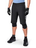 Alpinestars A-Dura Elite Shorts, zwarte MTB-mountainbikeshorts, gedragen met een lichtblauw shirt en zwarte kniebescherming, voorzien van een stevige drukknoopsluiting en een zak met rits, gecombineerd met zwarte fietsschoenen en handschoenen voor een complete off-road outfit.