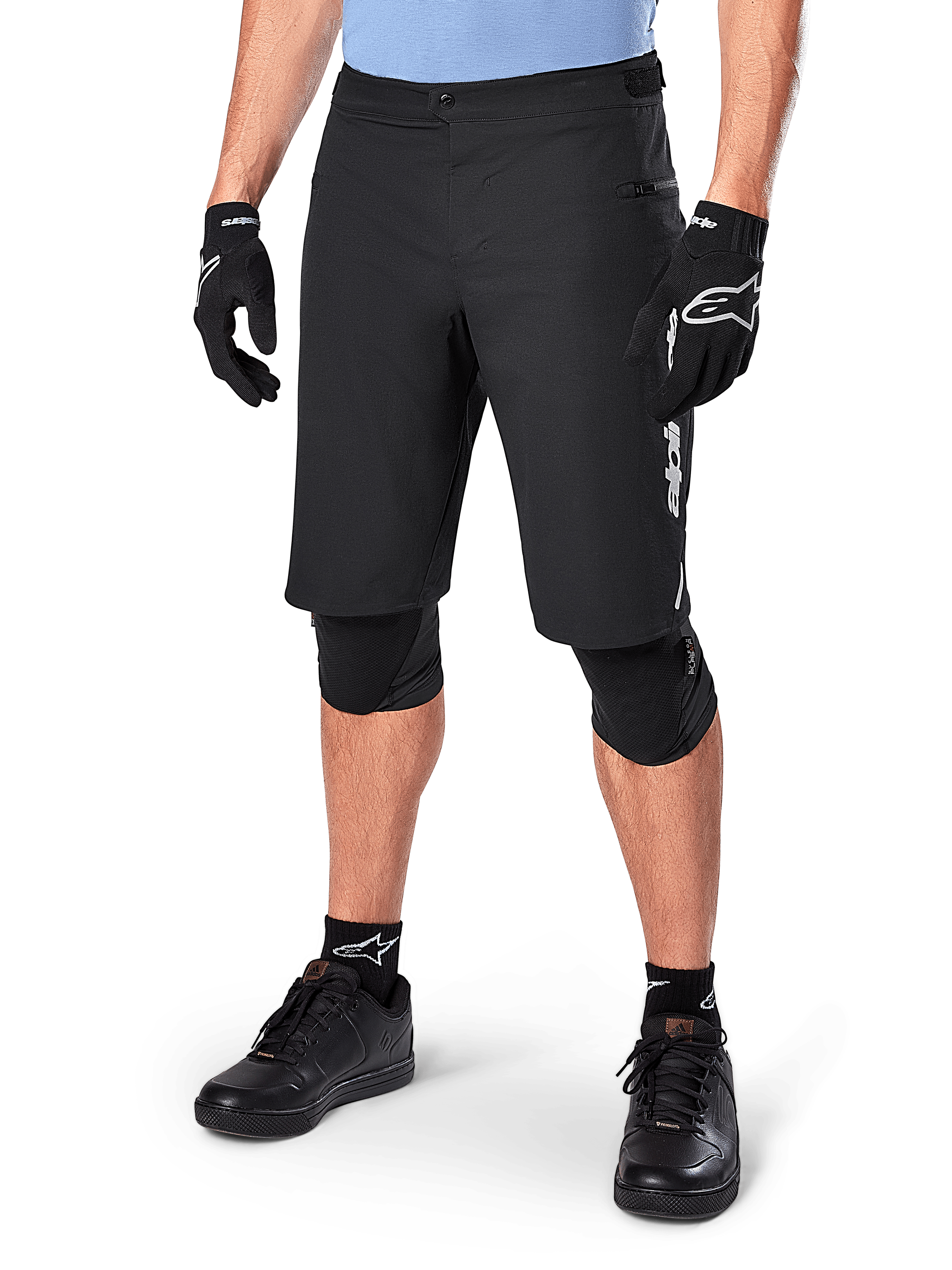Alpinestars A-Dura Elite Shorts, zwarte MTB-mountainbikeshorts, gedragen met een lichtblauw shirt en zwarte kniebescherming, voorzien van een stevige drukknoopsluiting en een zak met rits, gecombineerd met zwarte fietsschoenen en handschoenen voor een complete off-road outfit.