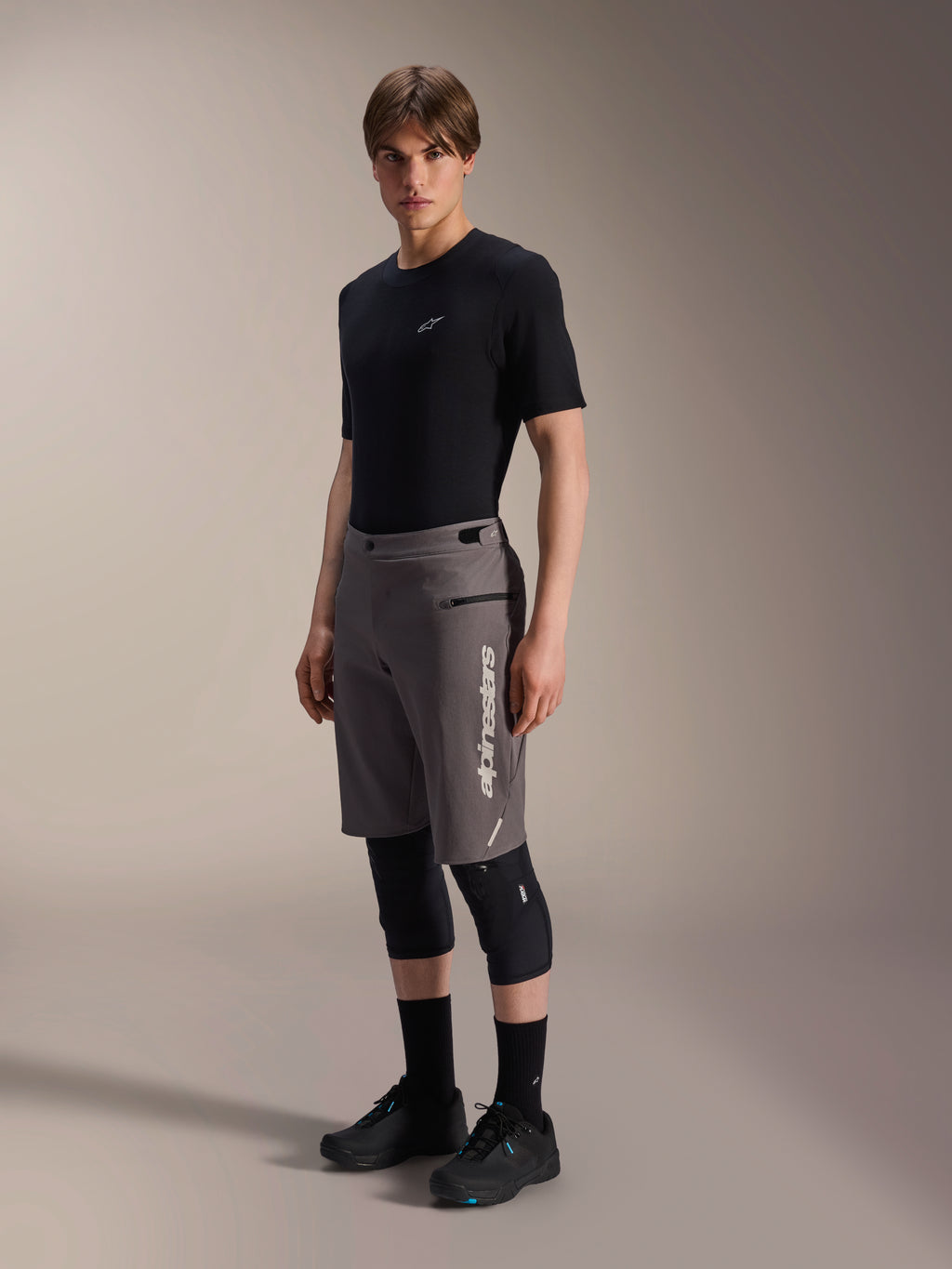 Alpinestars A-Dura Elite Shorts, donkergrijze mountainbike shorts, gedragen door een model met een zwart t-shirt en kniebeschermers, voorzien van een verticaal wit Alpinestars-logo op de linkerpijp, technisch Cordura-materiaal voor off-road fietsen