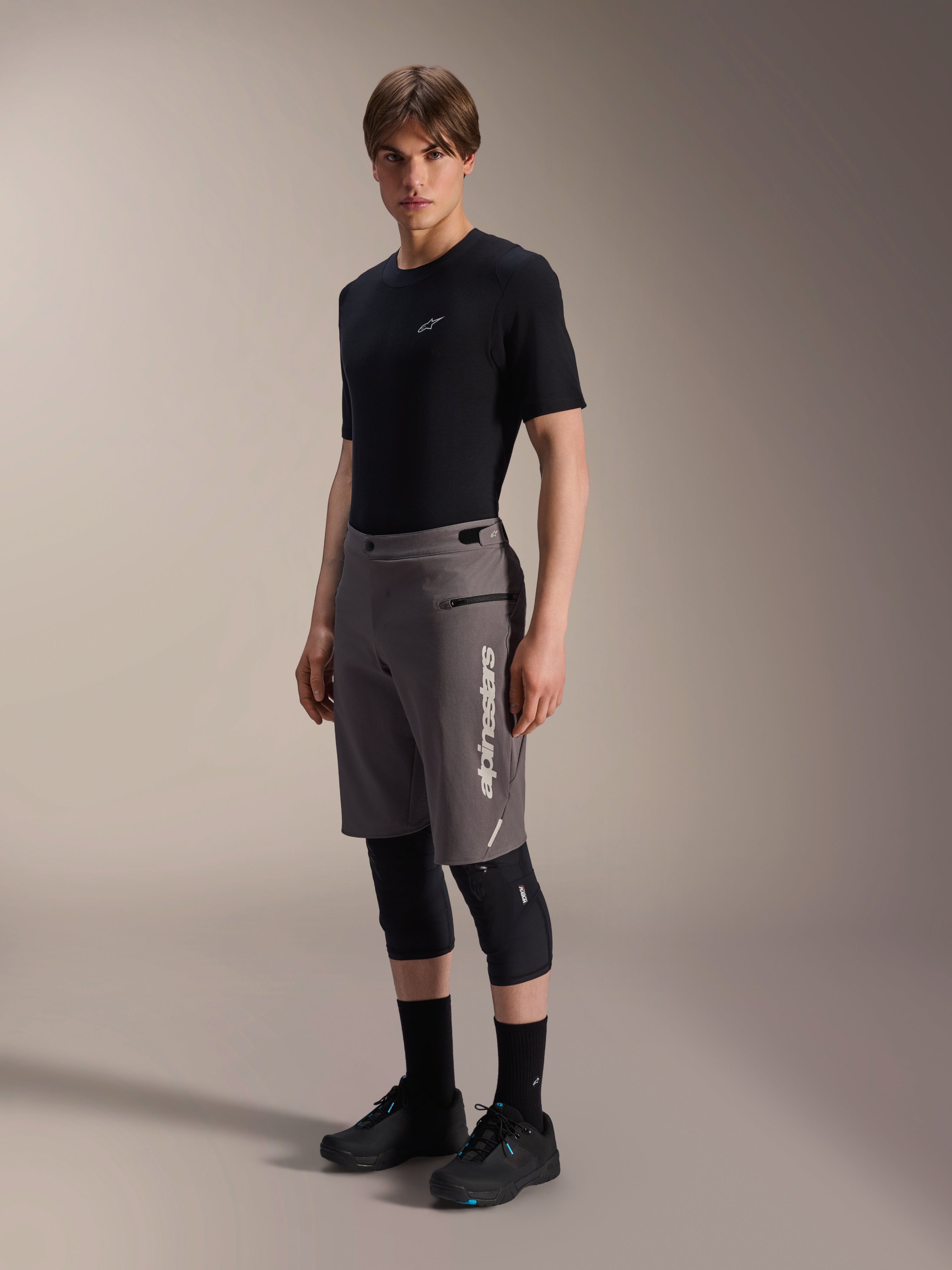 Alpinestars A-Dura Elite Shorts, donkergrijze mountainbike shorts, gedragen door een model met een zwart t-shirt en kniebeschermers, voorzien van een verticaal wit Alpinestars-logo op de linkerpijp, technisch Cordura-materiaal voor off-road fietsen
