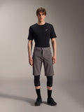 Alpinestars A-Dura Elite Shorts, donkergrijze mountainbikebroek, gedragen met een zwart T-shirt met ronde hals en zwarte kniebeschermers, knielange technische fietskleding met een drukknoopsluiting en duurzame Cordura-stof