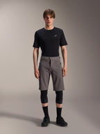 Alpinestars A-Dura Elite Shorts, donkergrijze mountainbikebroek, gedragen met een zwart T-shirt met ronde hals en zwarte kniebeschermers, knielange technische fietskleding met een drukknoopsluiting en duurzame Cordura-stof