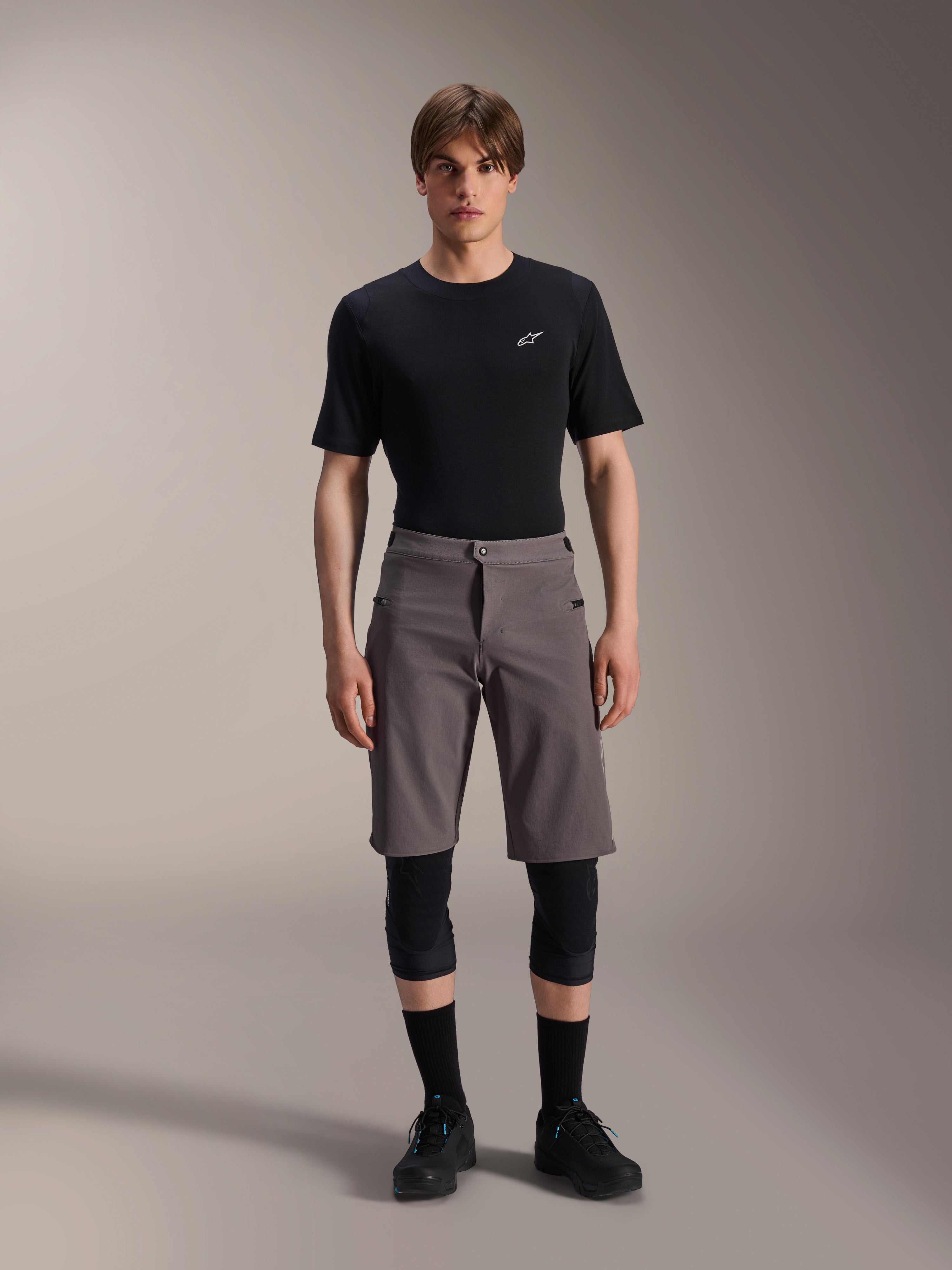 Alpinestars A-Dura Elite Shorts, donkergrijze mountainbikebroek, gedragen met een zwart T-shirt met ronde hals en zwarte kniebeschermers, knielange technische fietskleding met een drukknoopsluiting en duurzame Cordura-stof