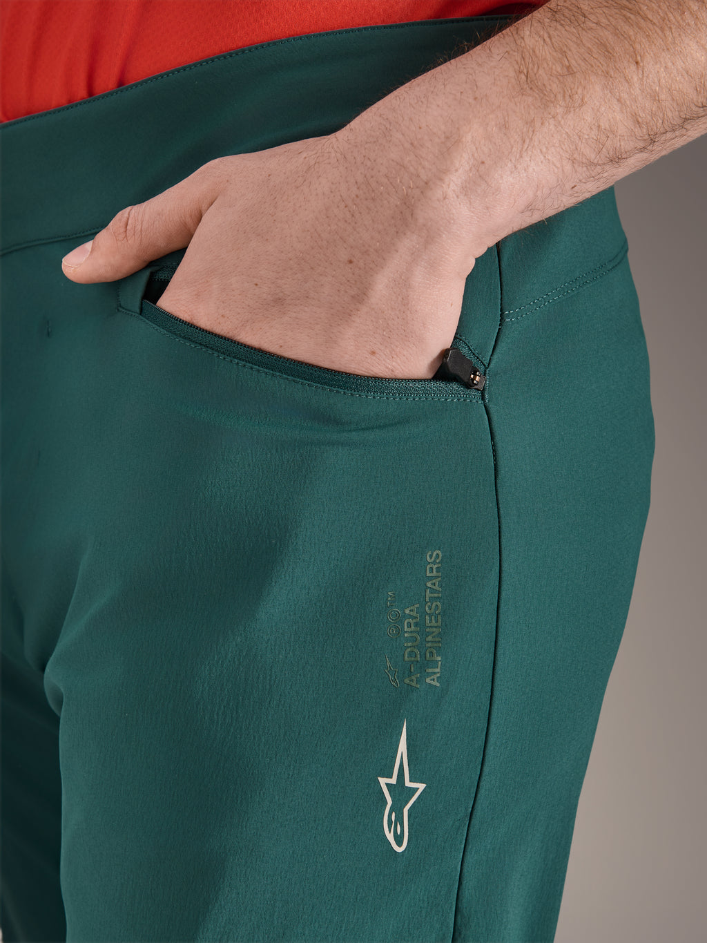 Alpinestars A-Dura Pants in Dark Pine groen, close-up van zak met ritssluiting en hand, voorzien van wit Alpinestars-logo en tekstbranding op het bovenbeen, duurzame MTB-fietsbroek