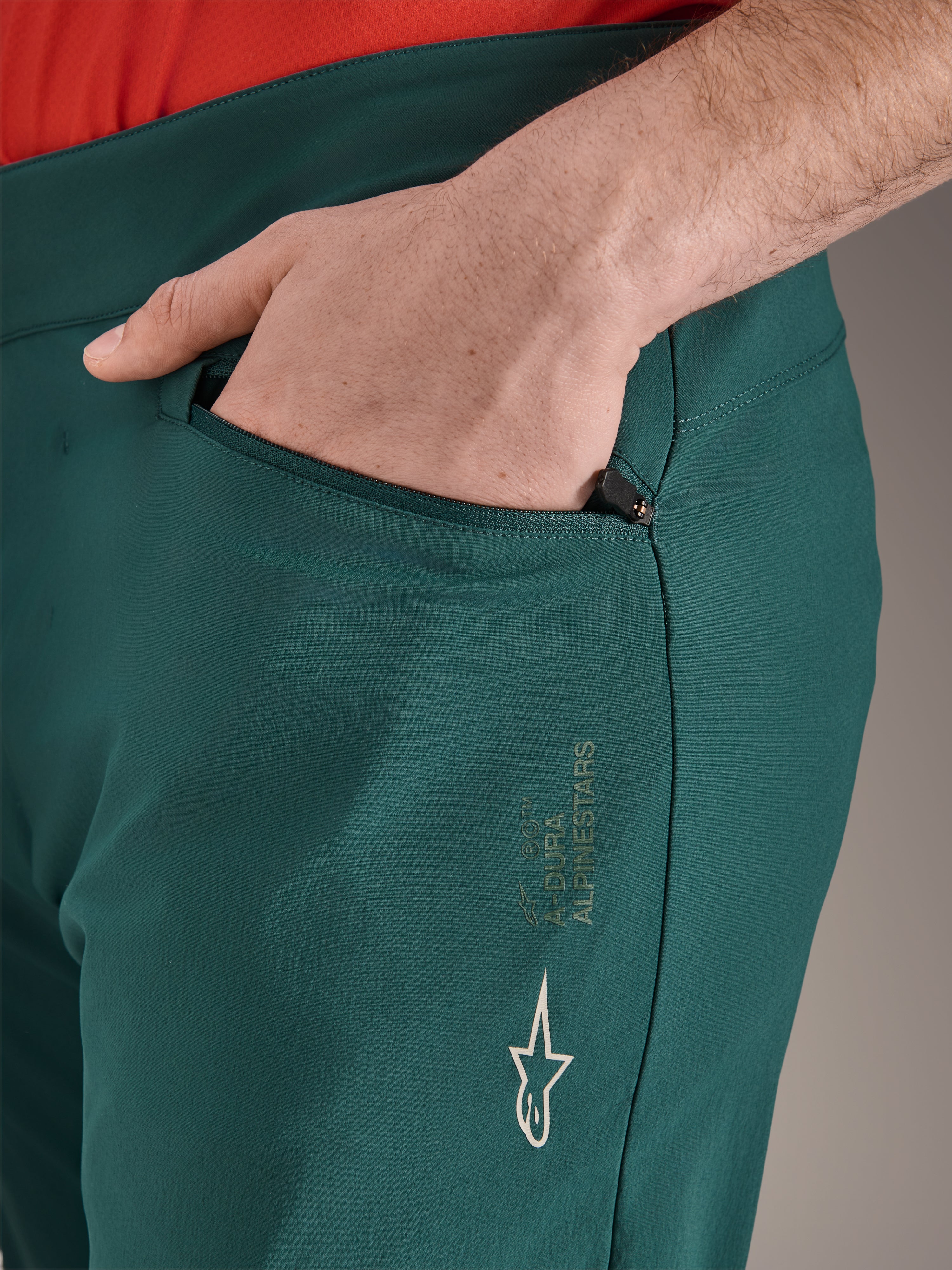 Alpinestars A-Dura Pants in Dark Pine groen, close-up van zak met ritssluiting en hand, voorzien van wit Alpinestars-logo en tekstbranding op het bovenbeen, duurzame MTB-fietsbroek
