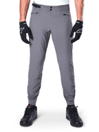 Alpinestars A-Dura Pants, donkergrijze mountainbikebroek, vooraanzicht gedragen met zwarte handschoenen en zwarte sneakers, met dubbele drukknoopsluiting bij de taille, handzakken met rits en elastische manchetten, duurzame fietskleding voor trailrijden