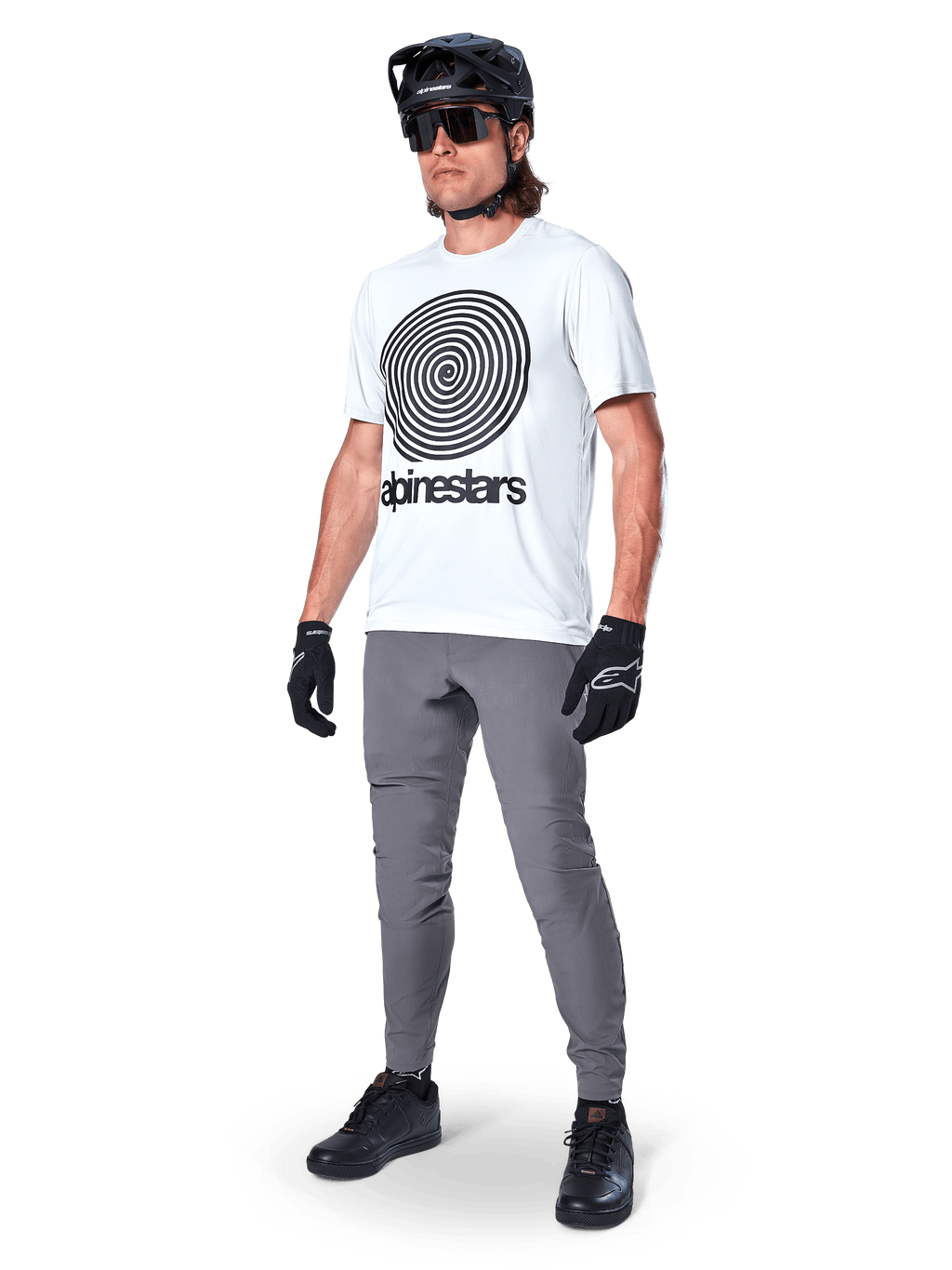 Alpinestars A-Dura broek, MTB-broek, donkergrijs, gedragen met wit T-shirt met spiraalprint, zwarte MTB-helm, zonnebril en zwarte handschoenen, duurzame 2-way stretch stof voor off-road fietsen