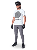 Alpinestars A-Dura broek, MTB-broek, donkergrijs, gedragen met wit T-shirt met spiraalprint, zwarte MTB-helm, zonnebril en zwarte handschoenen, duurzame 2-way stretch stof voor off-road fietsen