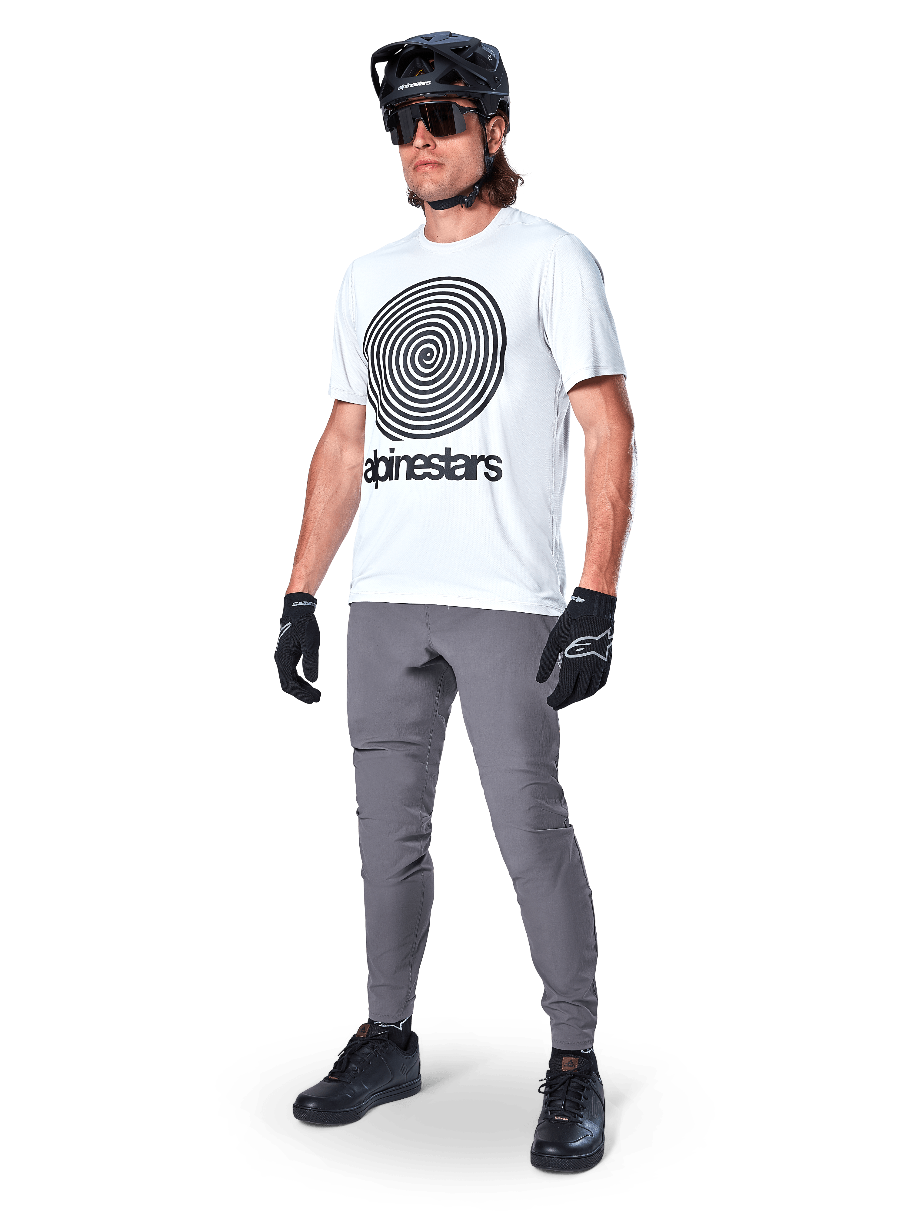 Alpinestars A-Dura broek, MTB-broek, donkergrijs, gedragen met wit T-shirt met spiraalprint, zwarte MTB-helm, zonnebril en zwarte handschoenen, duurzame 2-way stretch stof voor off-road fietsen