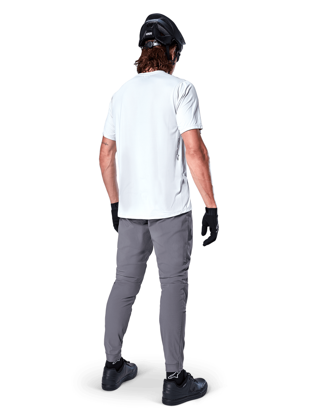 Alpinestars A-Dura broek, donkergrijze mountainbikebroek van achteren gezien, gedragen met een wit T-shirt met korte mouwen, zwarte MTB-helm, zwarte handschoenen en schoenen, regular fit ontwerp met elastische manchetten, duurzame off-road fietskleding
