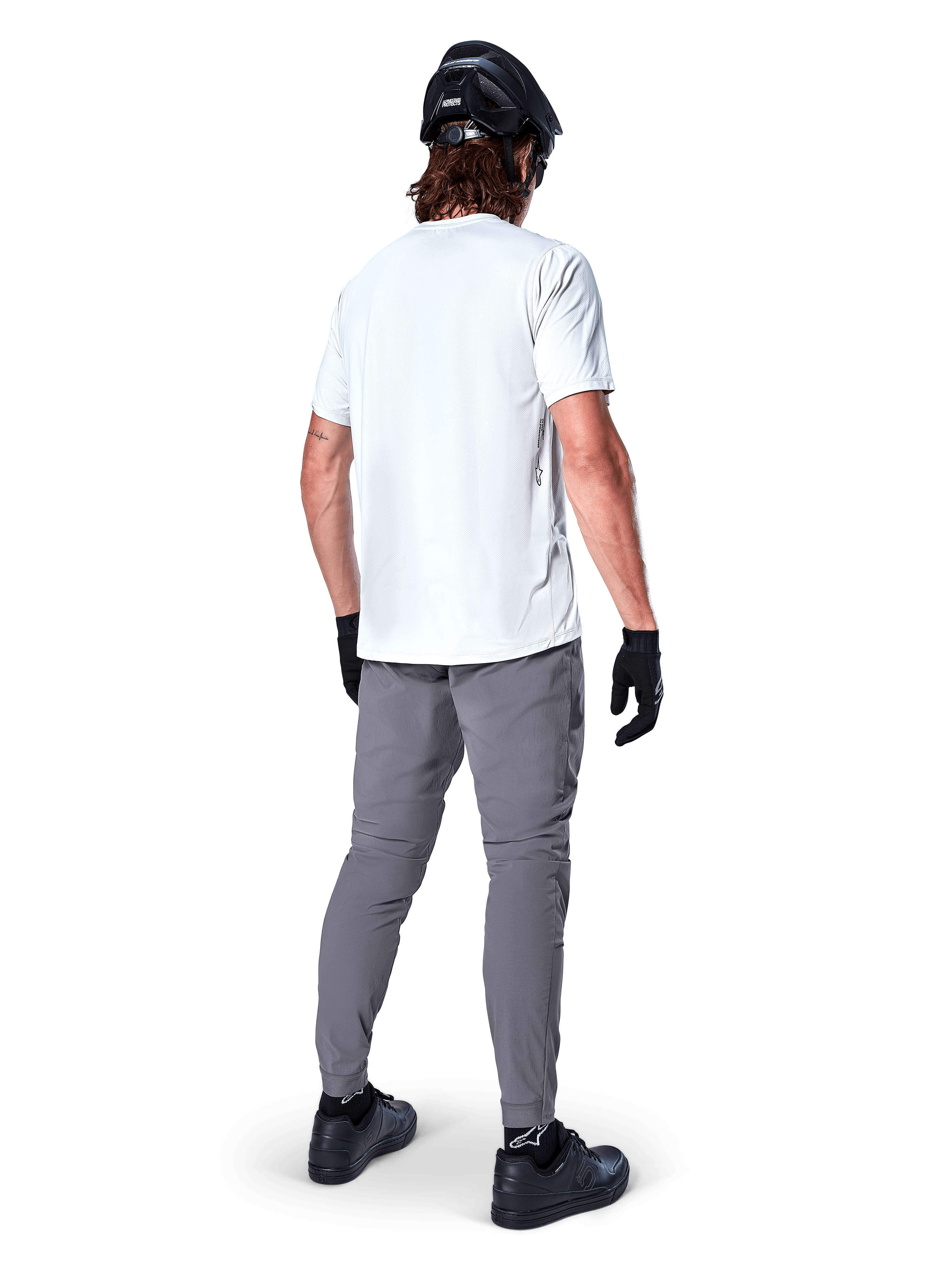 Alpinestars A-Dura broek, donkergrijze mountainbikebroek van achteren gezien, gedragen met een wit T-shirt met korte mouwen, zwarte MTB-helm, zwarte handschoenen en schoenen, regular fit ontwerp met elastische manchetten, duurzame off-road fietskleding
