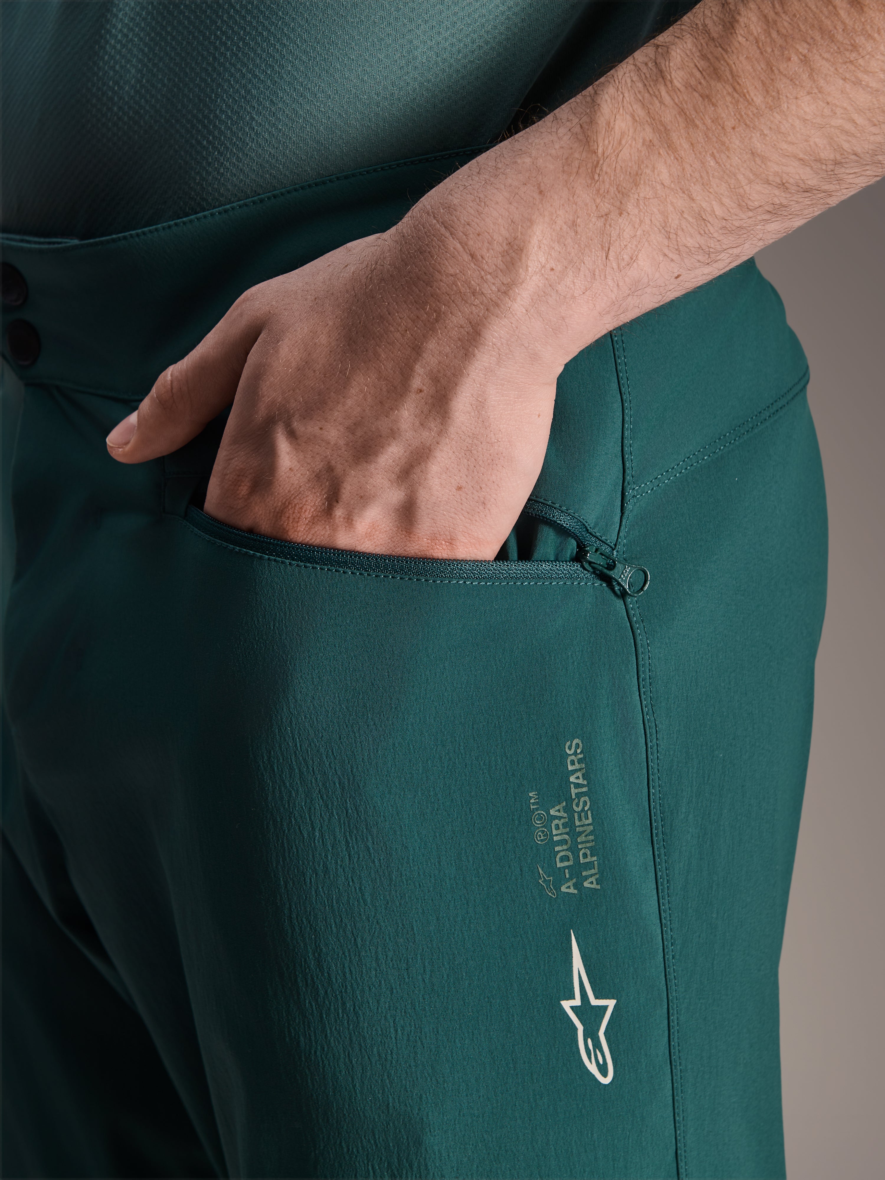 Alpinestars A-Dura Shorts in Dark Pine, detailopname van een hand in de zak met ritssluiting en mesh-voering, voorzien van wit Alpinestars-sterlogo en A-Dura-branding, duurzame textielconstructie voor mountainbiken