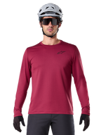 Mannelijke fietser in Alpinestars A-Dura Thermal Jersey, MTB-shirt met lange mouwen, bordeauxrood, gecombineerd met een grijze helm en zwarte zonnebril, voorzien van een klein zwart sterlogo op de borst, geïsoleerde fleece-stof voor mountainbiken in koud weer