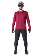 Alpinestars A-Dura Thermal Jersey in Burgundy, geïsoleerd fietsshirt met lange mouwen, mannelijk model met donkergrijze mountainbikebroek, witte helm, zwarte zonnebril en zwarte handschoenen, professionele MTB-uitrusting voor koud weer