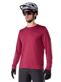 Alpinestars A-Dura Thermal Jersey, mountainbike-jersey met lange mouwen, bordeauxrood, voorzien van een geïsoleerde fleecevoering en klein zwart logo op de borst, gedragen met een grijze helm, zwarte zonnebril en zwarte handschoenen, ontworpen voor fietsen in koud weer