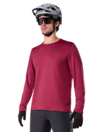 Alpinestars A-Dura Thermal Jersey, mountainbike-jersey met lange mouwen, bordeauxrood, voorzien van een geïsoleerde fleecevoering en klein zwart logo op de borst, gedragen met een grijze helm, zwarte zonnebril en zwarte handschoenen, ontworpen voor fietsen in koud weer