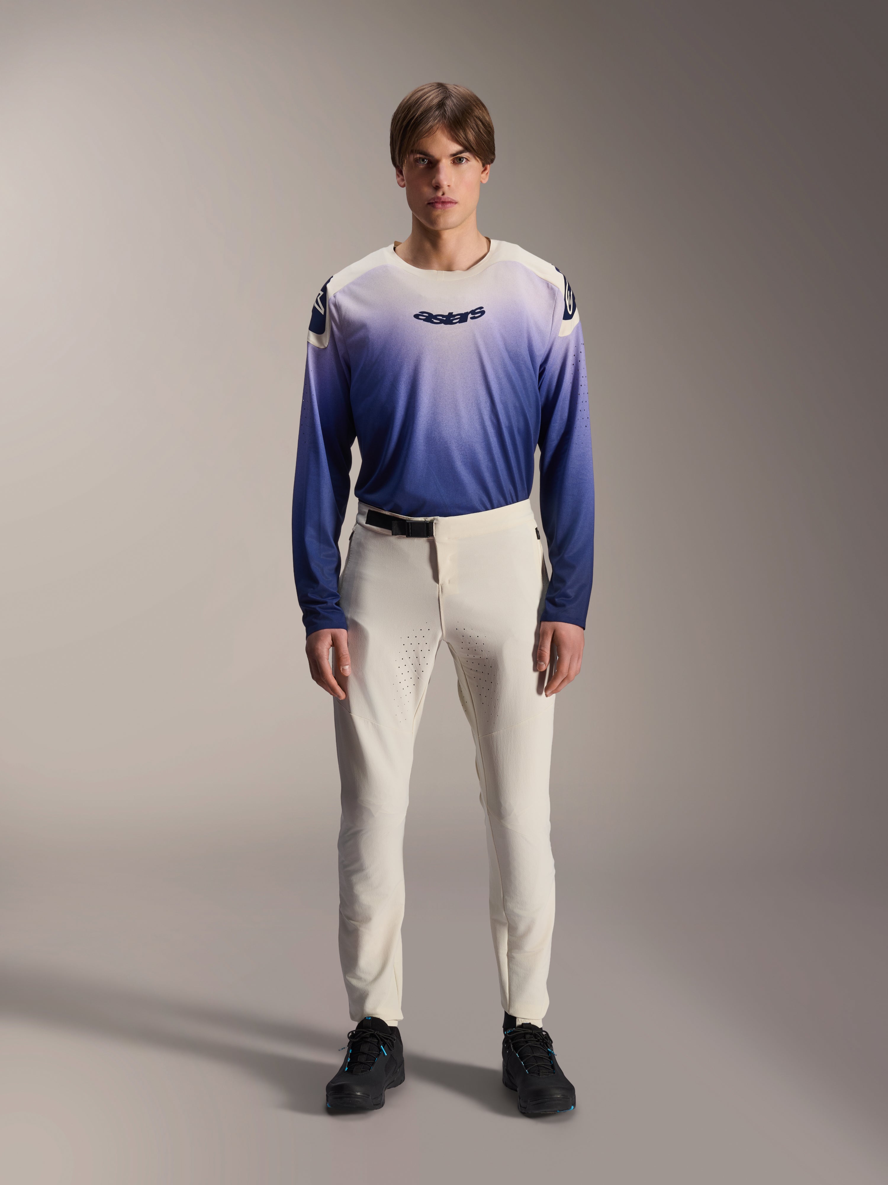 Alpinestars A-Aria Polartec Maker Elite Jersey, mountainbike-jersey met lange mouwen, Baja Blue en wit kleurverloop, voorzien van het astars-logo op de borst, gedragen door een model met een gebroken witte broek en zwarte schoenen, hoogwaardige fietskleding