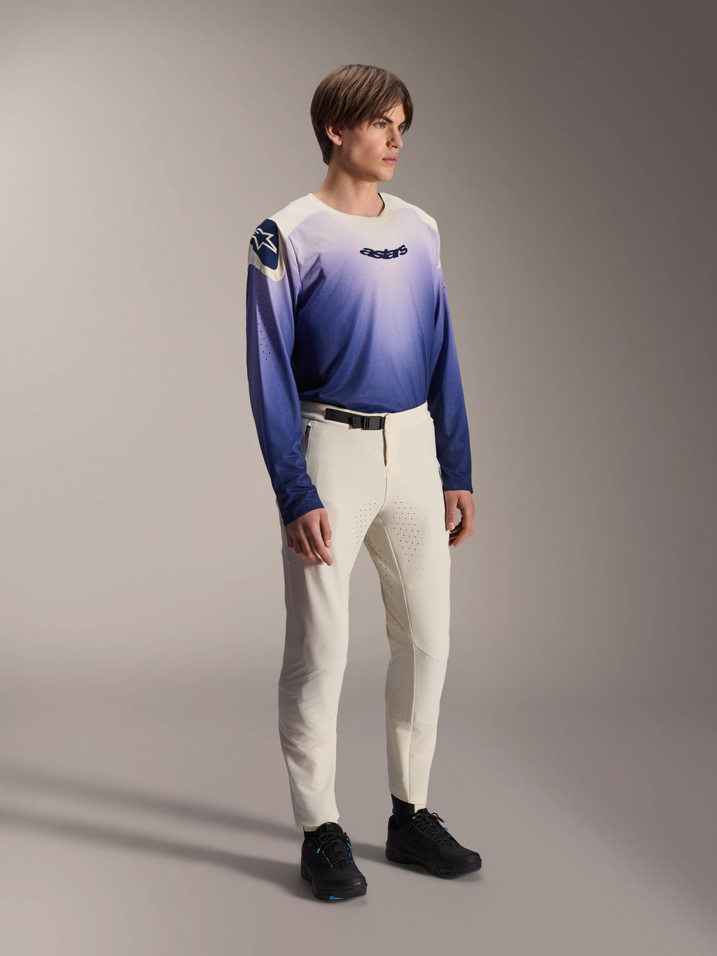 Mannelijk model draagt de Alpinestars A-Aria Polartec Maker Elite Jersey in Baja Blue, voorzien van een wit-blauw kleurverloop met een zwart 'astars'-logo op de borst, gecombineerd met een gebroken witte mountainbikebroek en zwarte schoenen, prestatiegerichte MTB-kleding voor trailrijden.