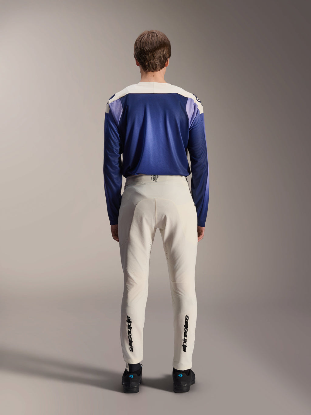 Alpinestars A-Aria Polartec Maker Elite Jersey met lange mouwen, Baja Blue en gebroken wit, achteraanzicht van model in technische mountainbike-uitrusting inclusief gebroken witte broek met Alpinestars-branding op de onderbenen, performance wieleroutfit voor off-road trails