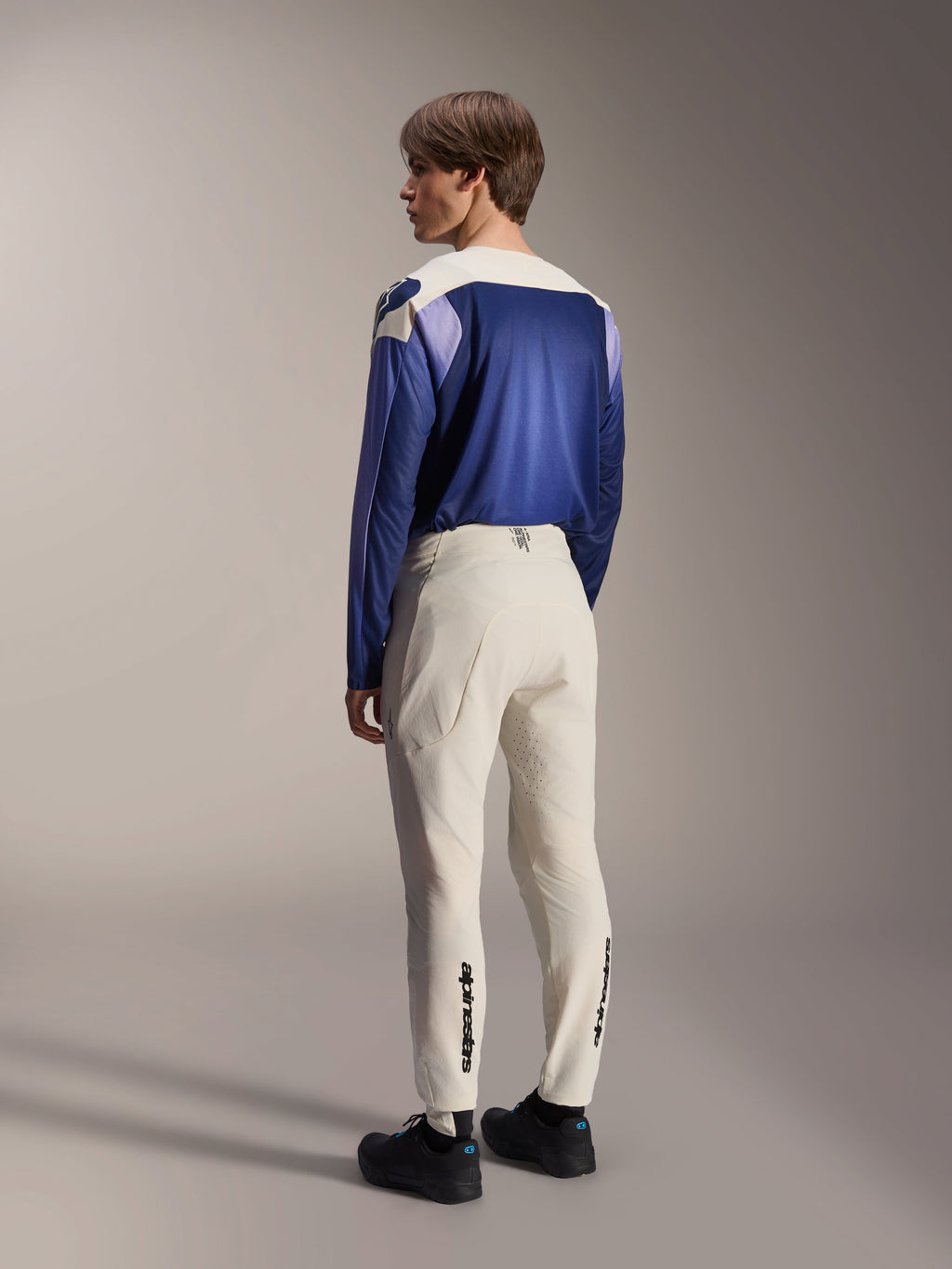 Alpinestars A-Aria Polartec Maker Elite jersey met lange mouwen, Baja Blue, mannelijk model van achteren gezien met gebroken witte MTB-broek en zwarte schoenen, blauw en wit kleurvlakontwerp, technische mountainbike-uitrusting